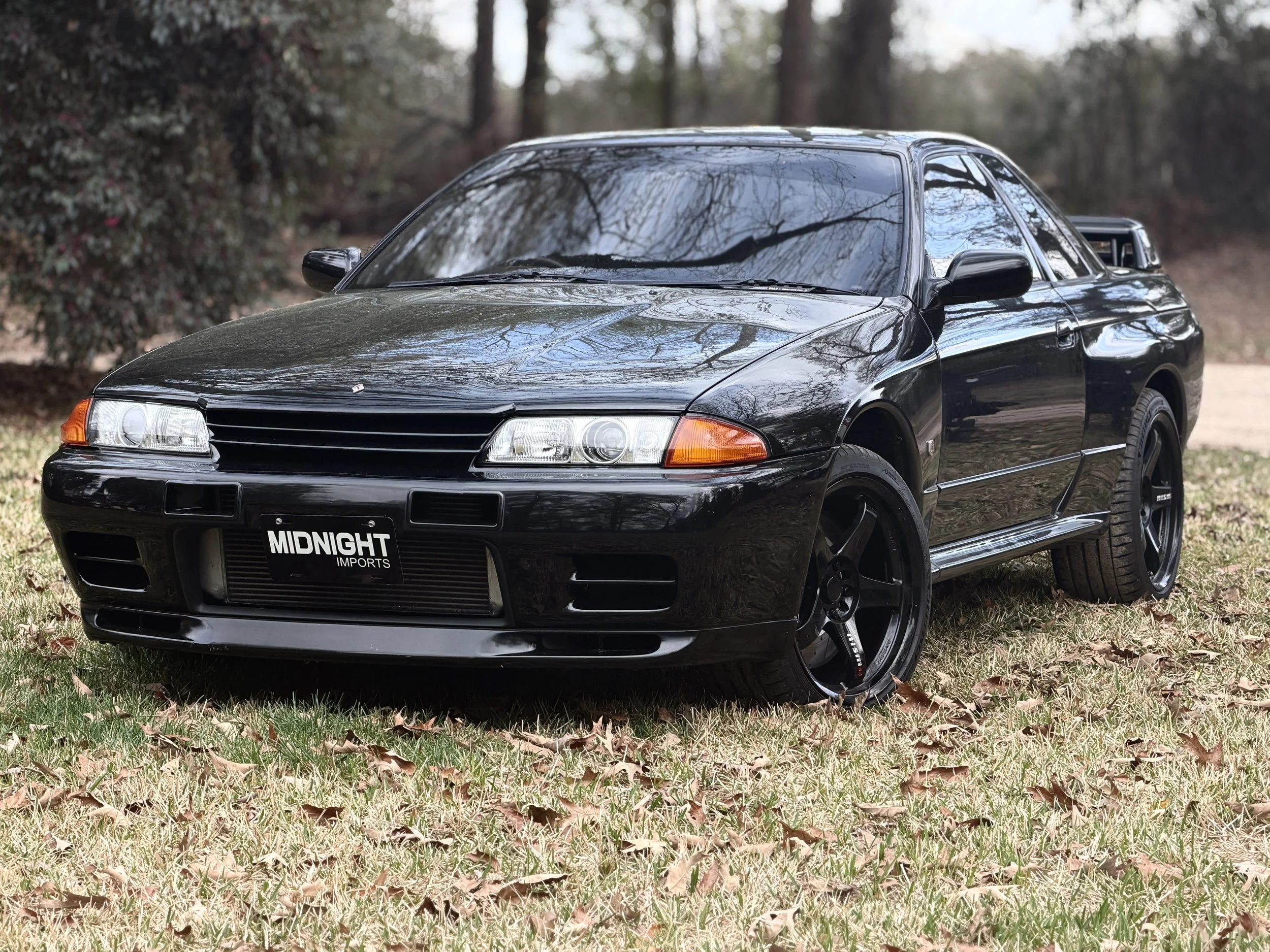 Black sports car parked on grass with trees in the background, featuring a sleek design with tinted windows and custom black wheels, displaying a license plate that reads 'MIDNIGHT IMPORTS'.