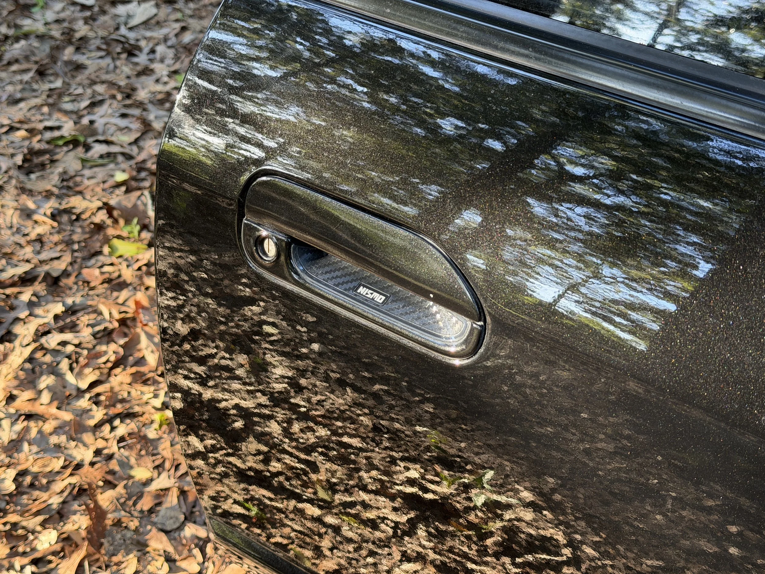 Close-up of a black car's headlight and front panel reflecting trees and the sky, with fallen leaves on the ground nearby.
