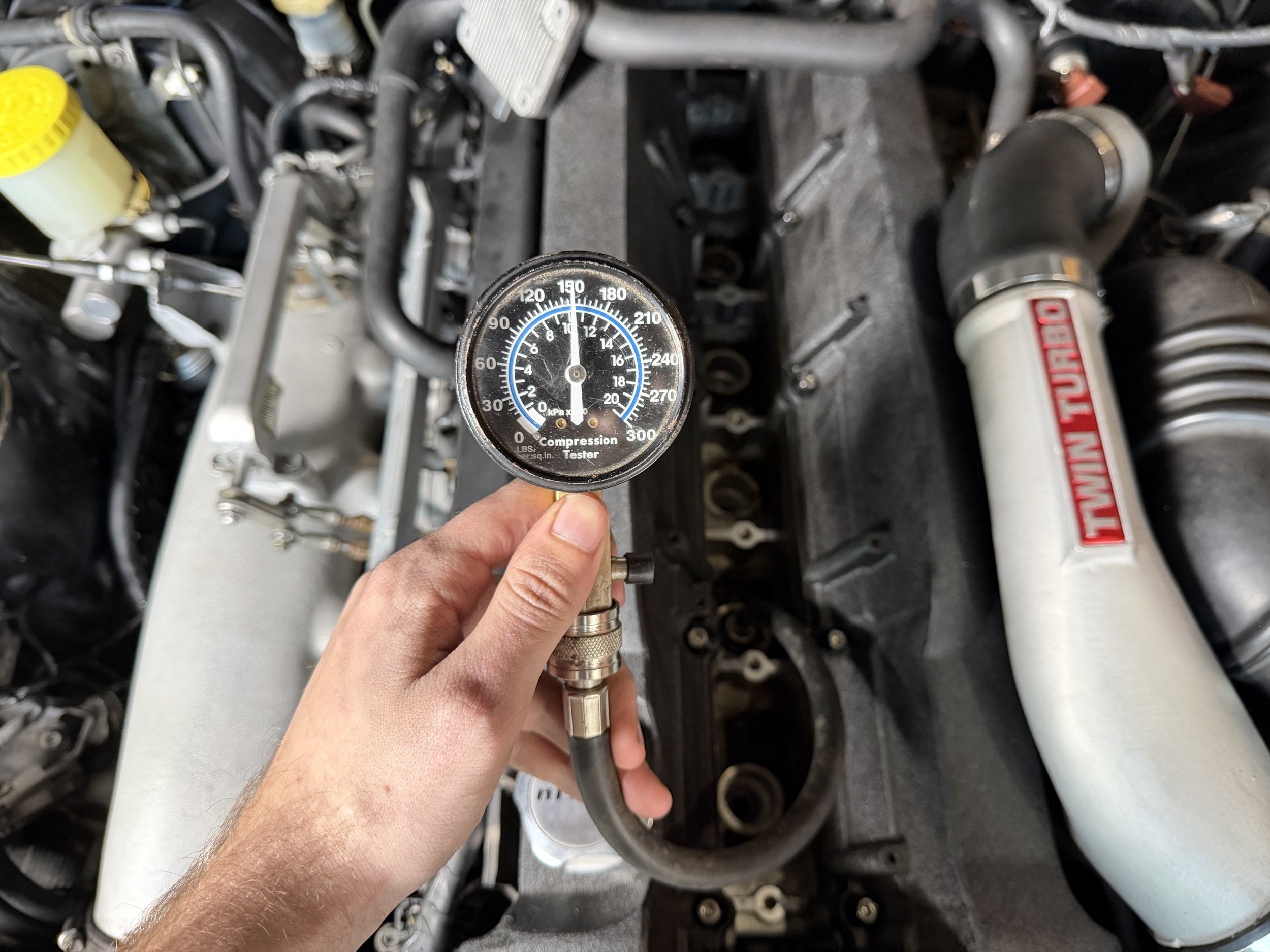 Close-up of a person holding a compression tester gauge above an engine with a Twin-Turbo label on the exhaust pipe, indicating a vehicle engine compression test.
