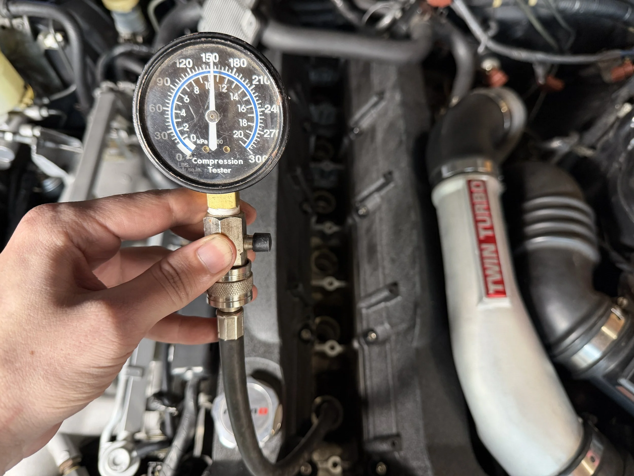 A person holding a compression tester gauge connected to a car engine for testing compression, with engine components visible in the background.