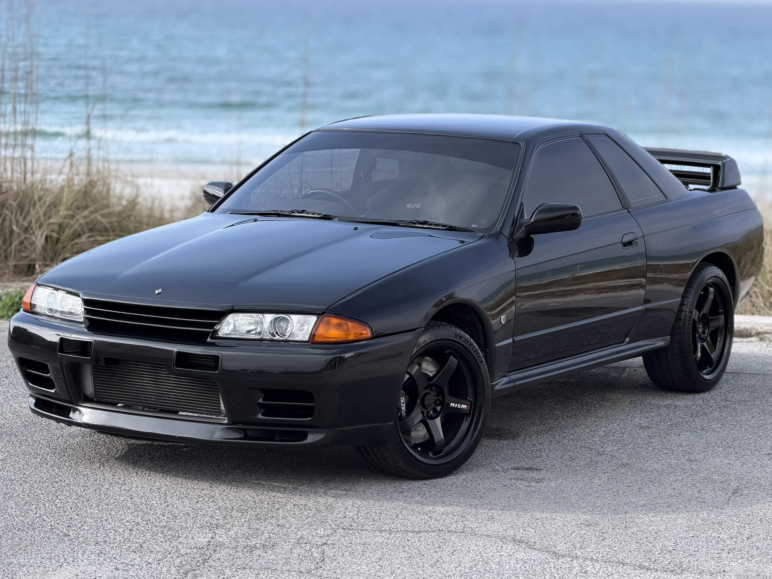 Black sports car parked near a beach with ocean and grass in the background.