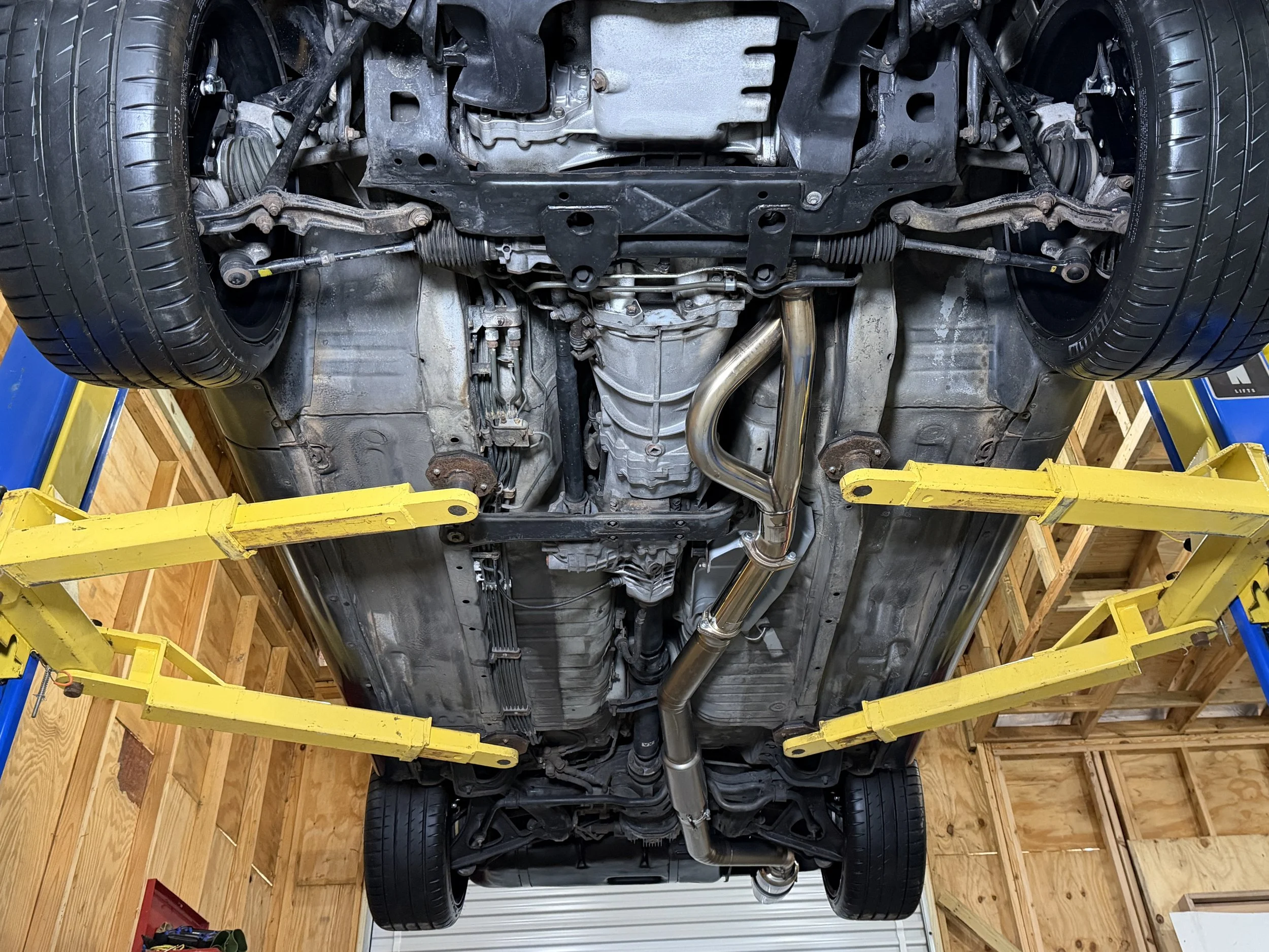 Underside of a car lifted on a yellow car lift in a garage, showing the exhaust system, suspension, and other undercarriage components.