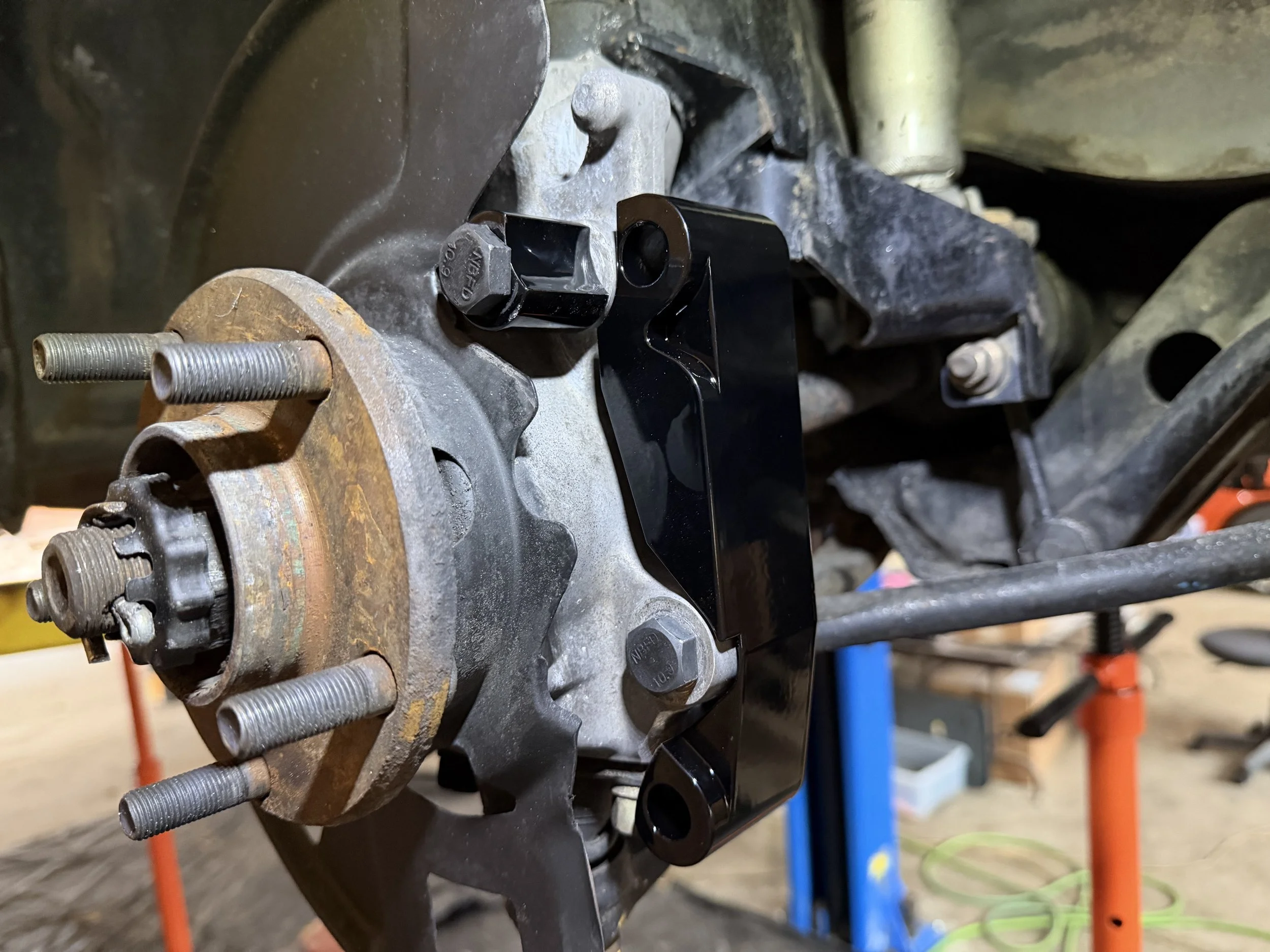 Close-up of a car wheel hub assembly with rusty rotor, black mounting bracket, and attached components, on a car in a garage or workshop.
