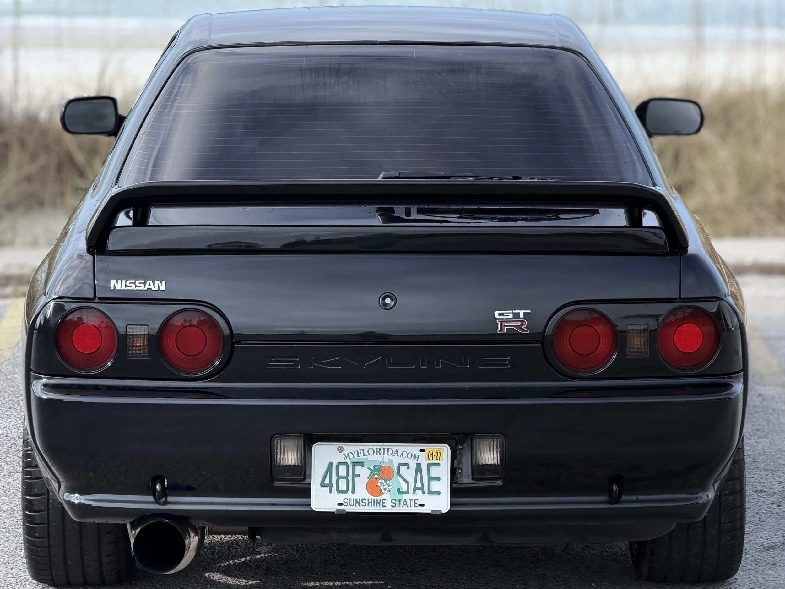 Rear view of a black Nissan Skyline GT-R car with a spoiler, round taillights, a visible exhaust pipe, and a Florida license plate reading 48FSAE, parked on the side of a road with blurred background.