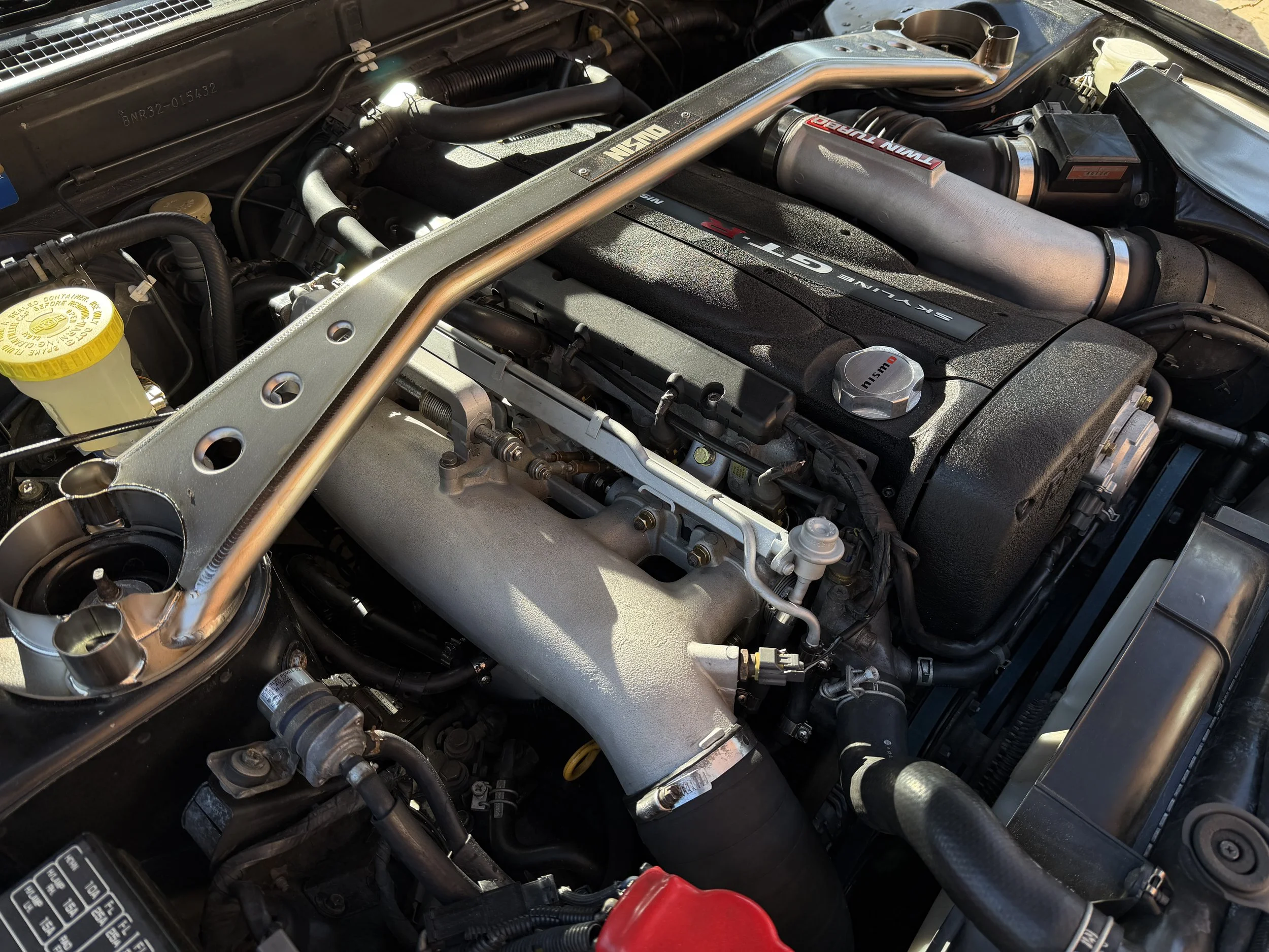 A close-up view of a car engine bay, featuring a large silver strut bar and a black engine cover with "SEKUKEN GT-R" text, various hoses, and other engine components.