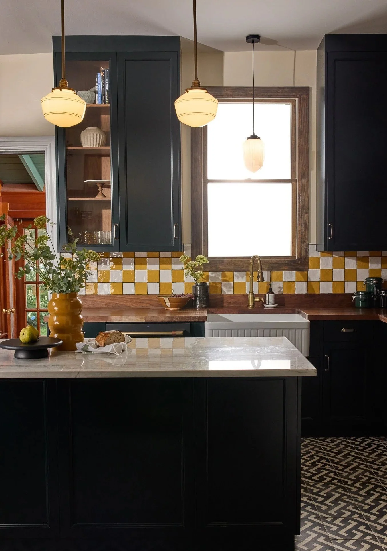 Kitchen with dark cabinets, yellow tile backsplash, and a large window. The countertop has a large yellow vase with green foliage, a black tray with a green apple, and bread. Two pendant lights hang above, and a patterned black and white floor is visible.