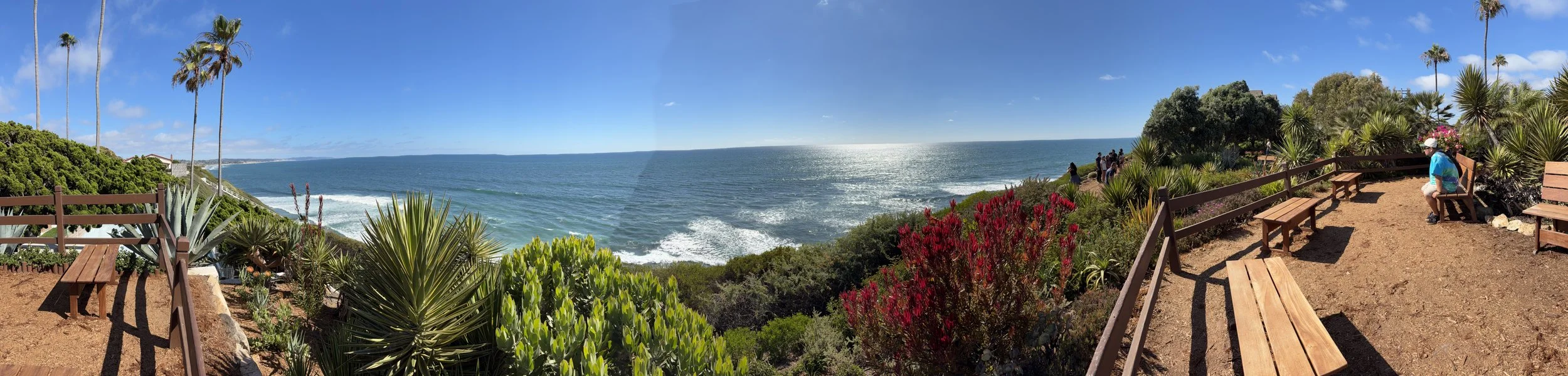Regarding IMG_1194.jpg, use this alt text:  Panoramic coastal park view with wooden benches along a fenced dirt path and lush plants overlooking the bright blue ocean.