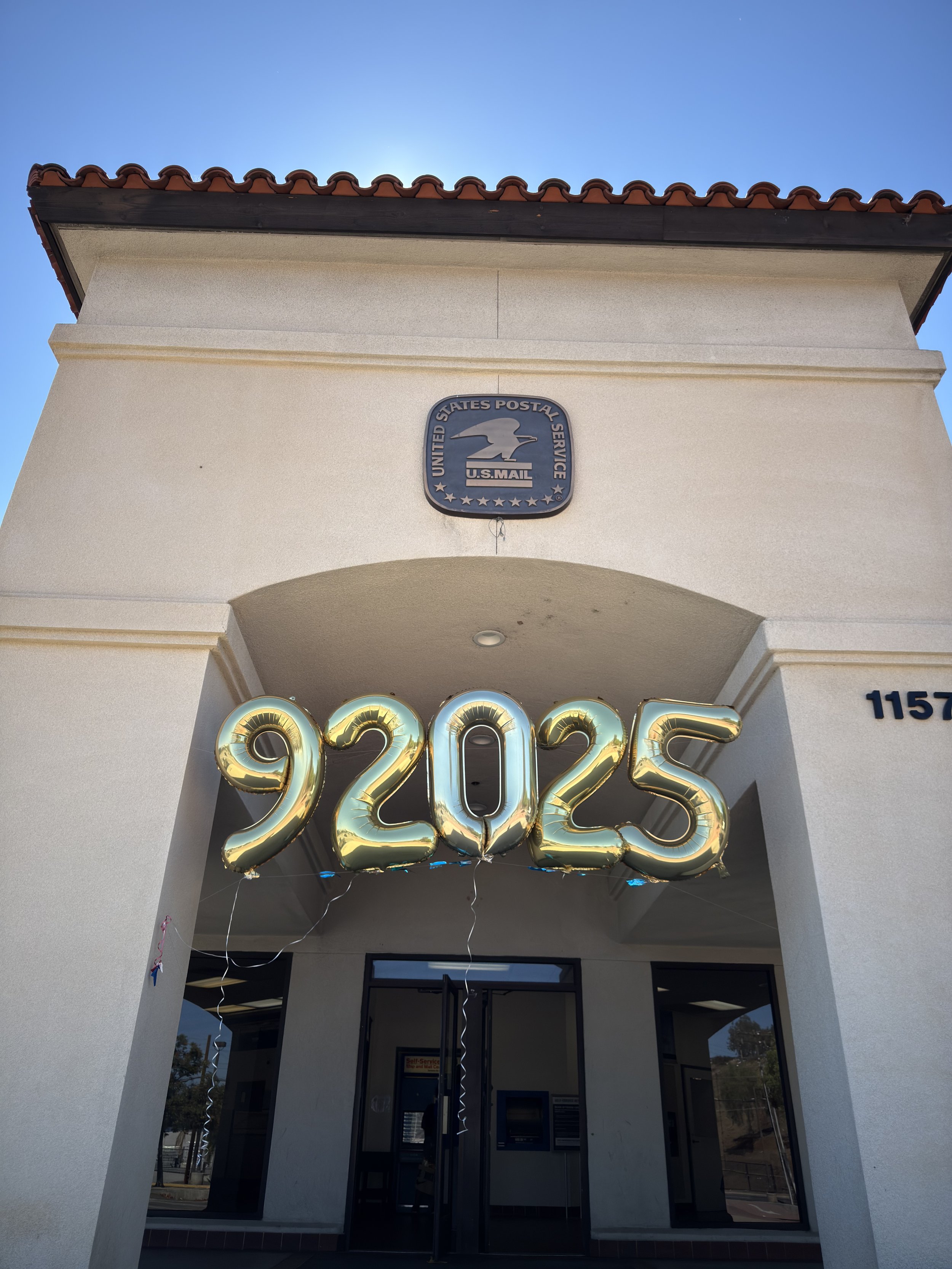 Large gold balloons spelling "92025" hang over the entrance of a United States Postal Service building in Escondido on a sunny day.