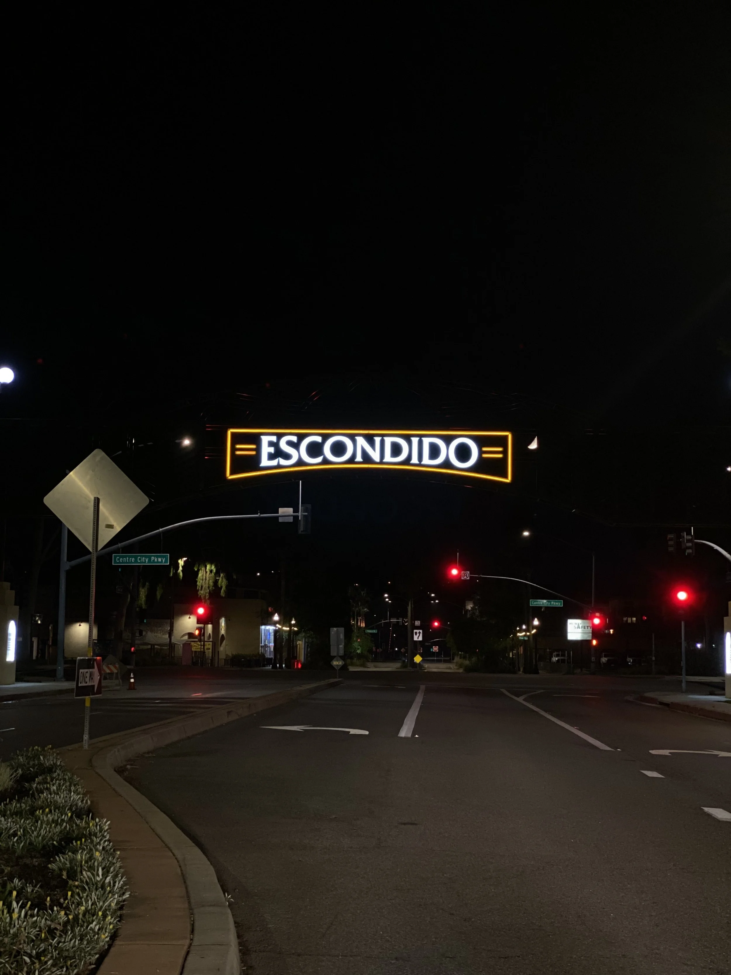 Nighttime view of Grand Avenue in Escondido, CA. A bright, illuminated archway sign reading "ESCONDIDO" spans across the empty street under red traffic lights.