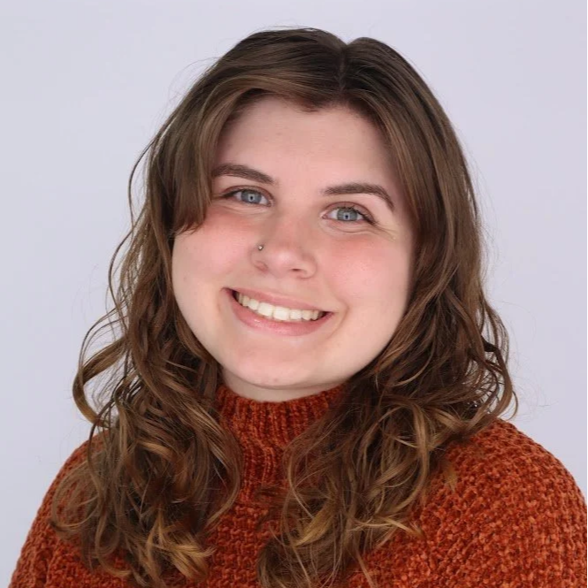 A young woman with wavy brown hair and blue eyes smiling, wearing a rust-colored sweater, and a nose piercing against a plain light gray background.