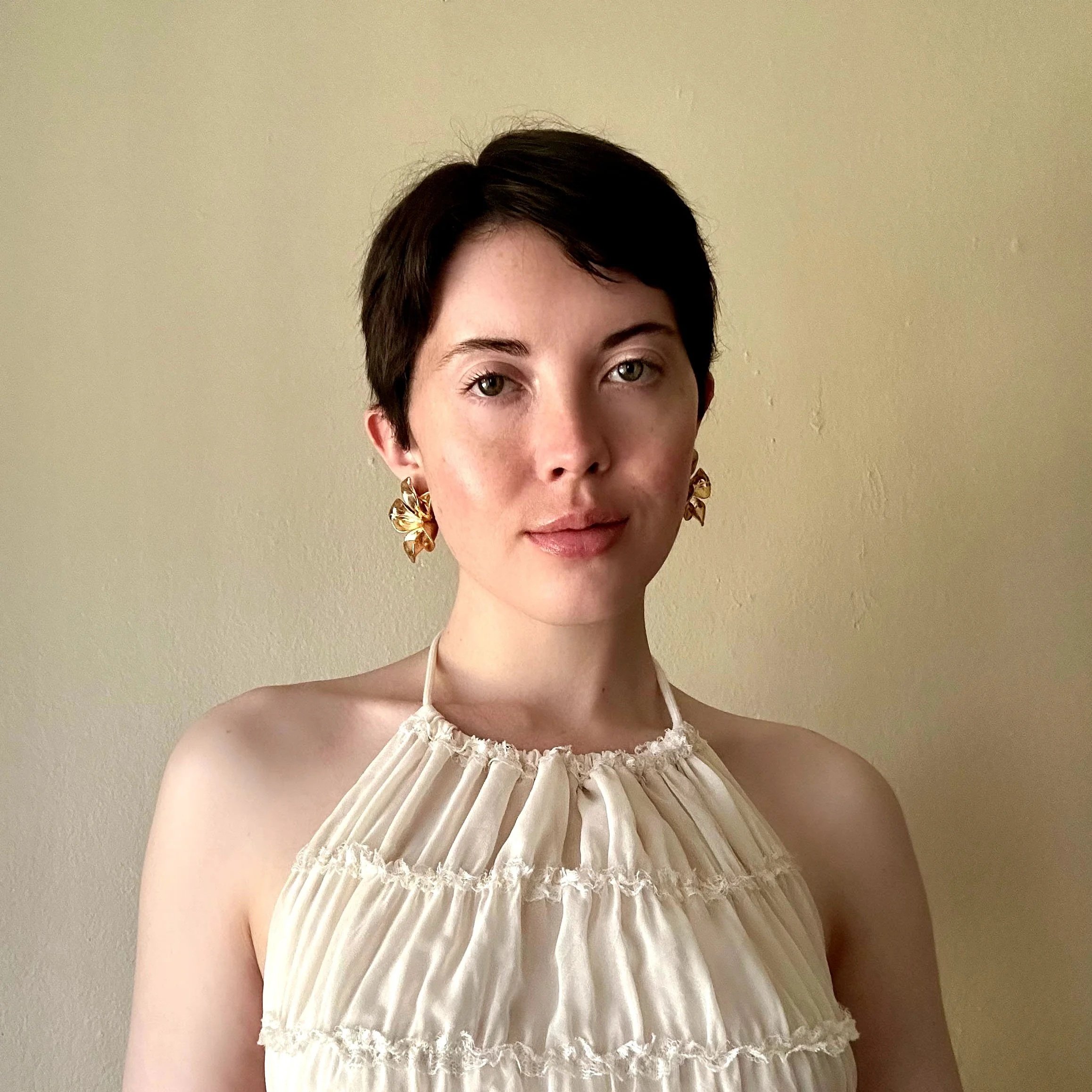 Close-up of a woman with short dark hair, wearing large gold earrings and a cream-colored dress with ruffles, standing against a plain beige wall.