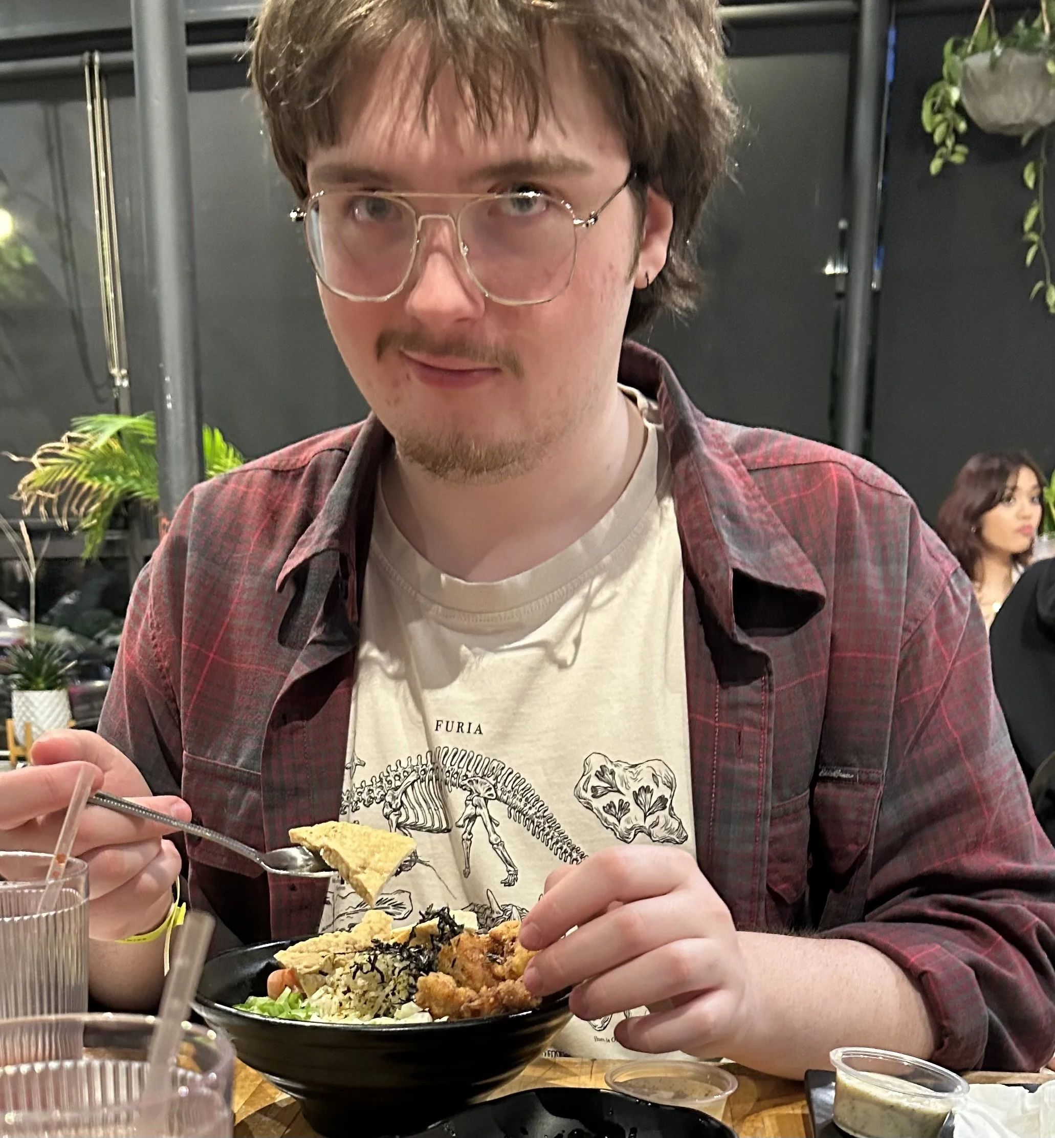 A young man with glasses and a beard eating from a black bowl filled with sushi, fried food, lettuce, and seaweed. He is wearing a white T-shirt with a dinosaur print underneath a red plaid shirt. There are drinks and soy sauce on the table. Two women are seated in the background.