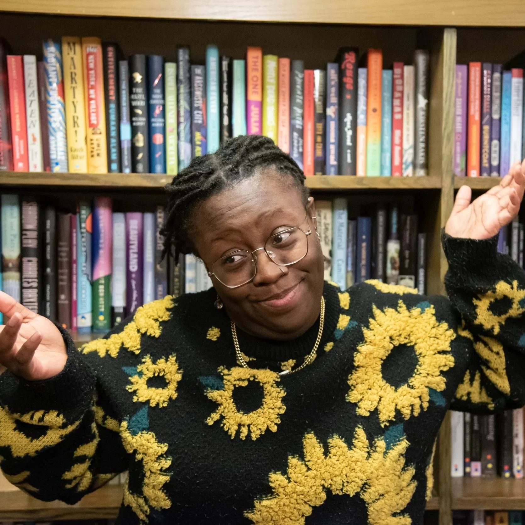 A woman with glasses and earrings in front of a bookshelf, smiling, and wearing a black sweater with yellow and blue flower patterns.