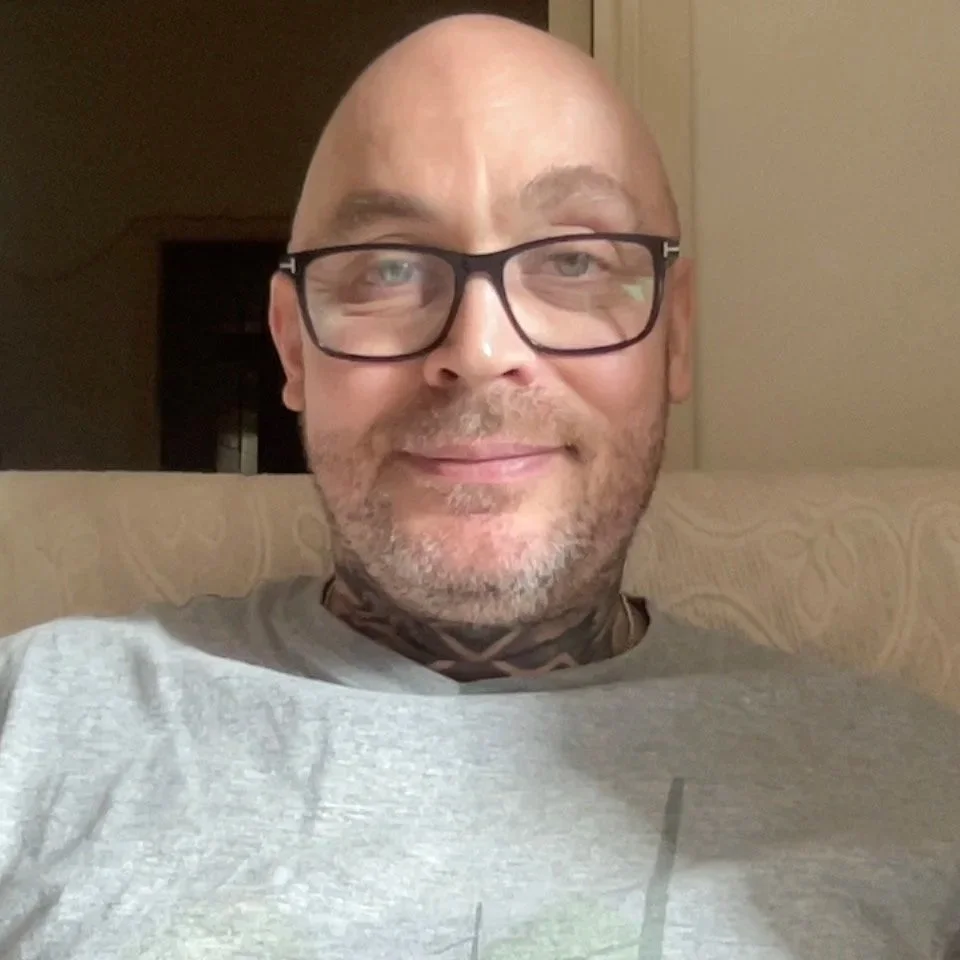 A man with glasses smiling, sitting on a beige patterned couch in an indoor setting.