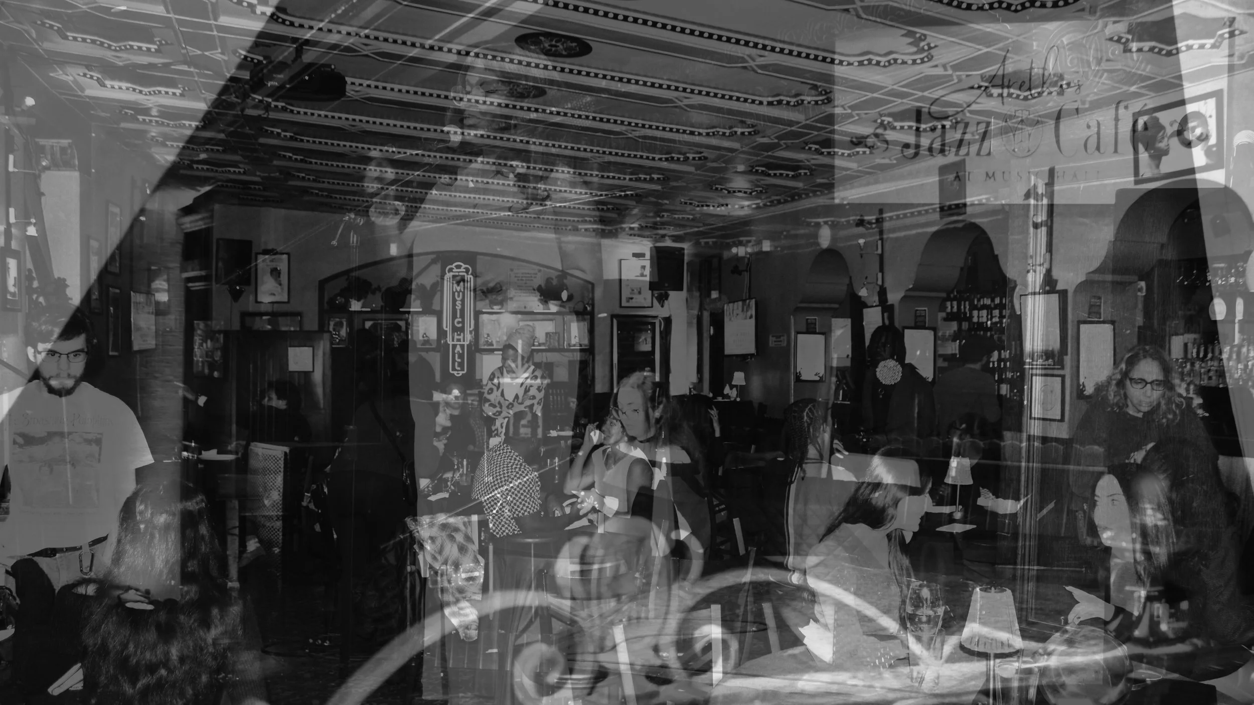 People sitting and standing in a lively indoor space with colorful decor, including a sign that says 'Jazz' and framed pictures on the walls, suggesting a cafe or jazz club atmosphere.