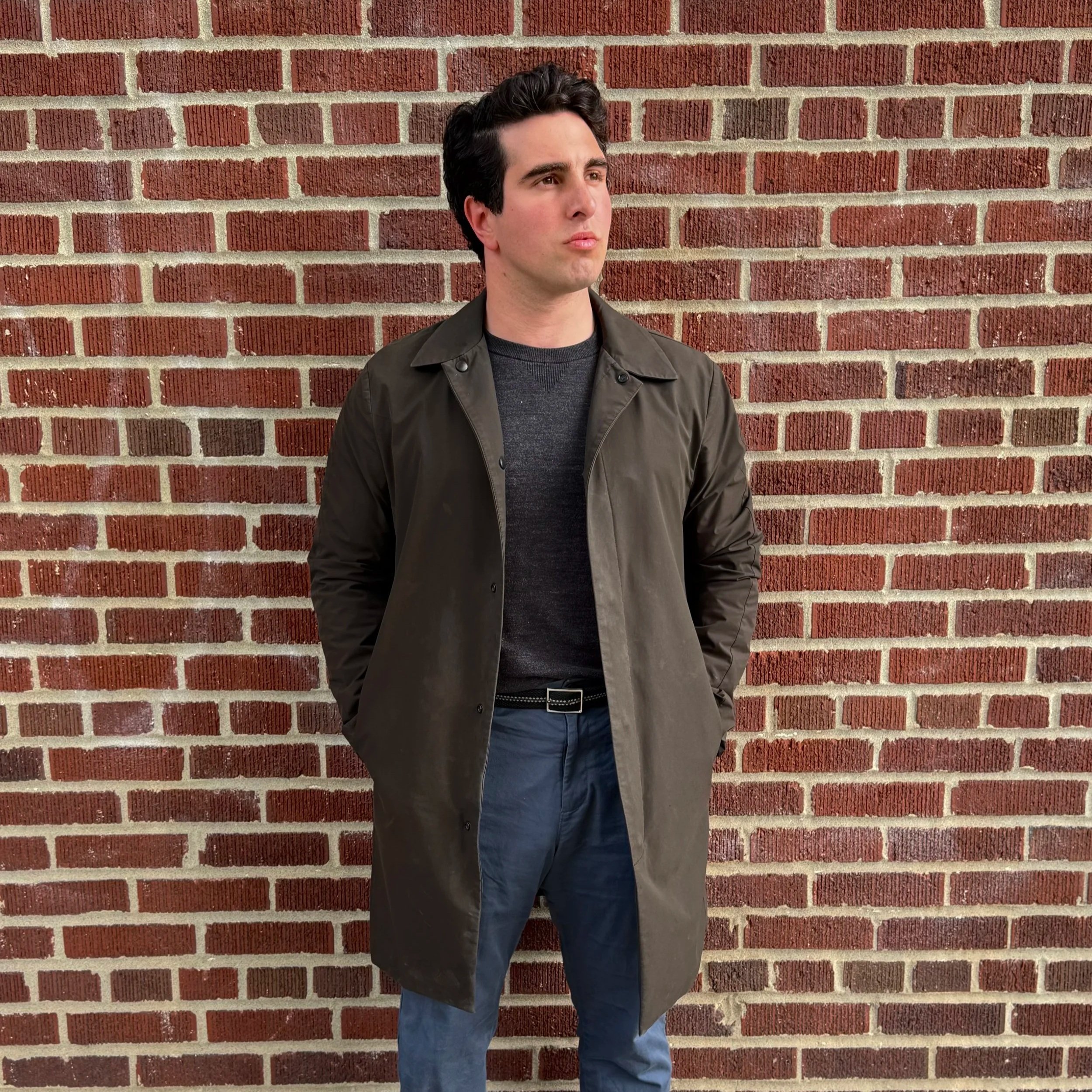A young man with dark hair, wearing a brown jacket and blue jeans, standing against a red brick wall, looking to his left with a serious expression.