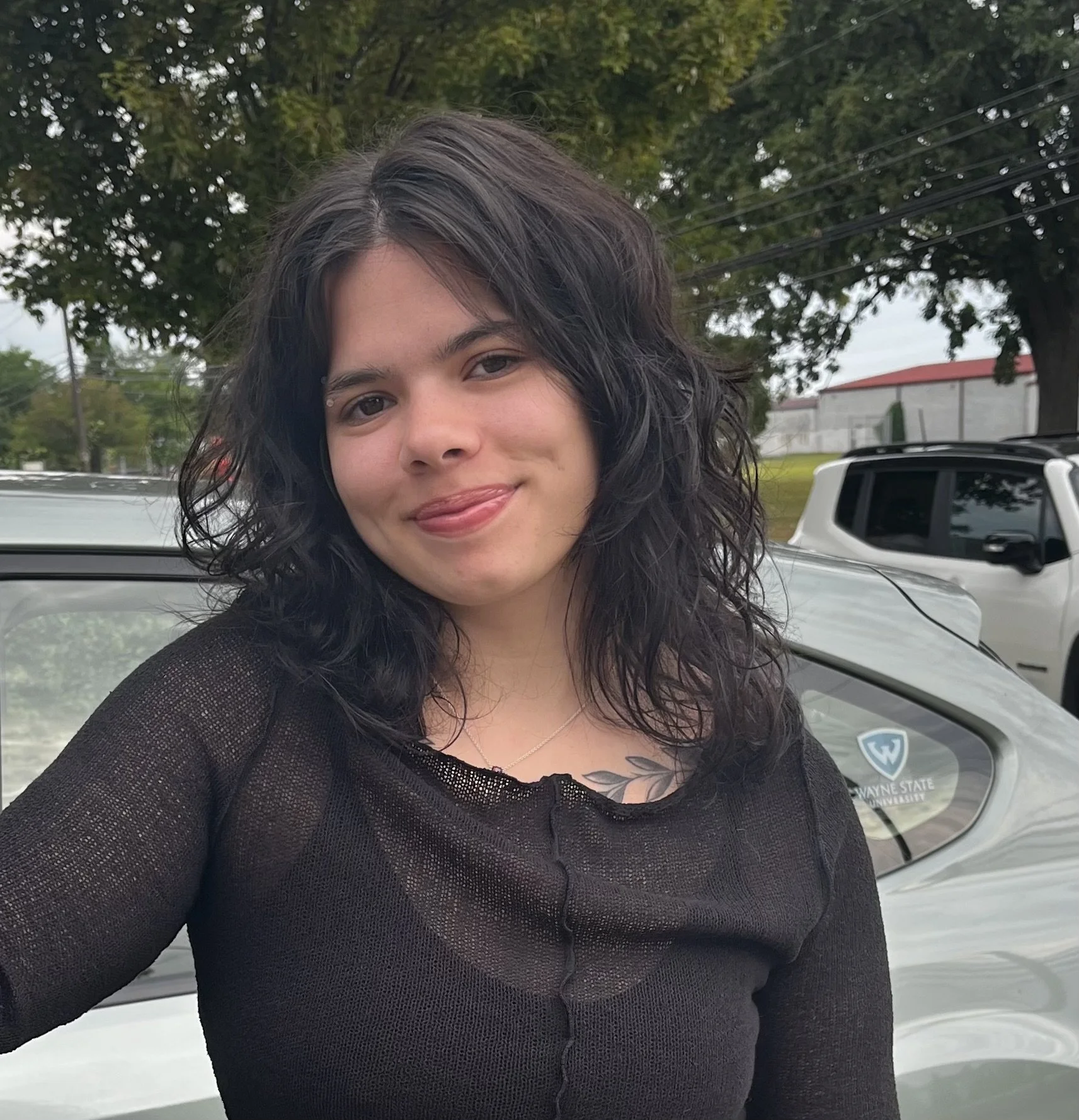 Close-up of a young woman with curly dark hair, smiling, wearing a black top, standing outdoors in front of a white vehicle with a Wayne State University sticker, trees, and a building in the background.