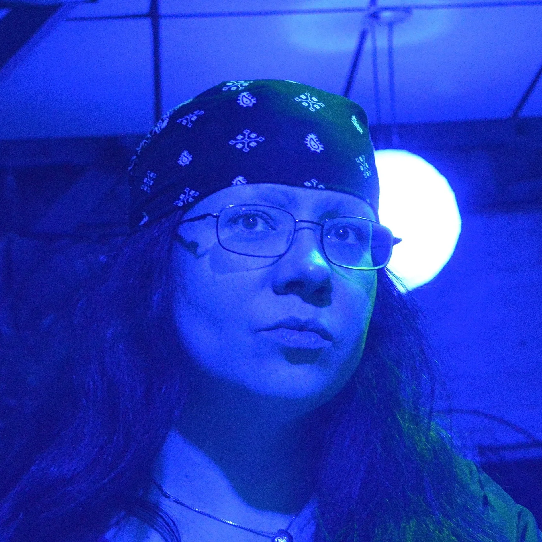 A person wearing glasses and a green bandana with white patterns, illuminated by blue light. The person has long hair and visible eyes.
