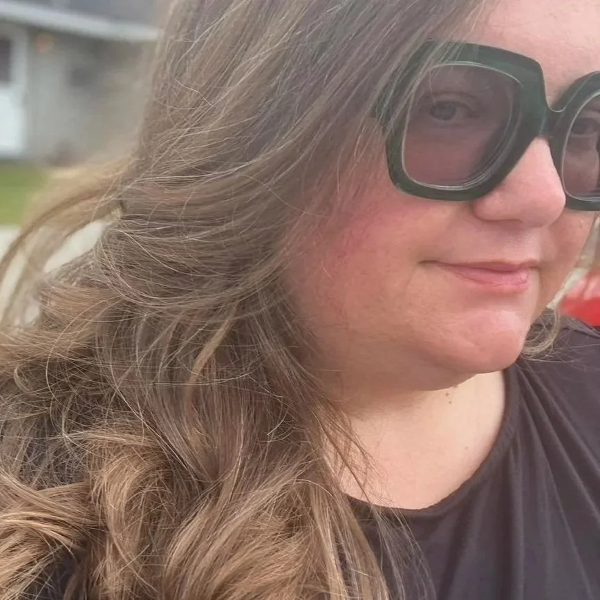 Close-up of a woman with long, wavy hair wearing large black glasses, smiling slightly.