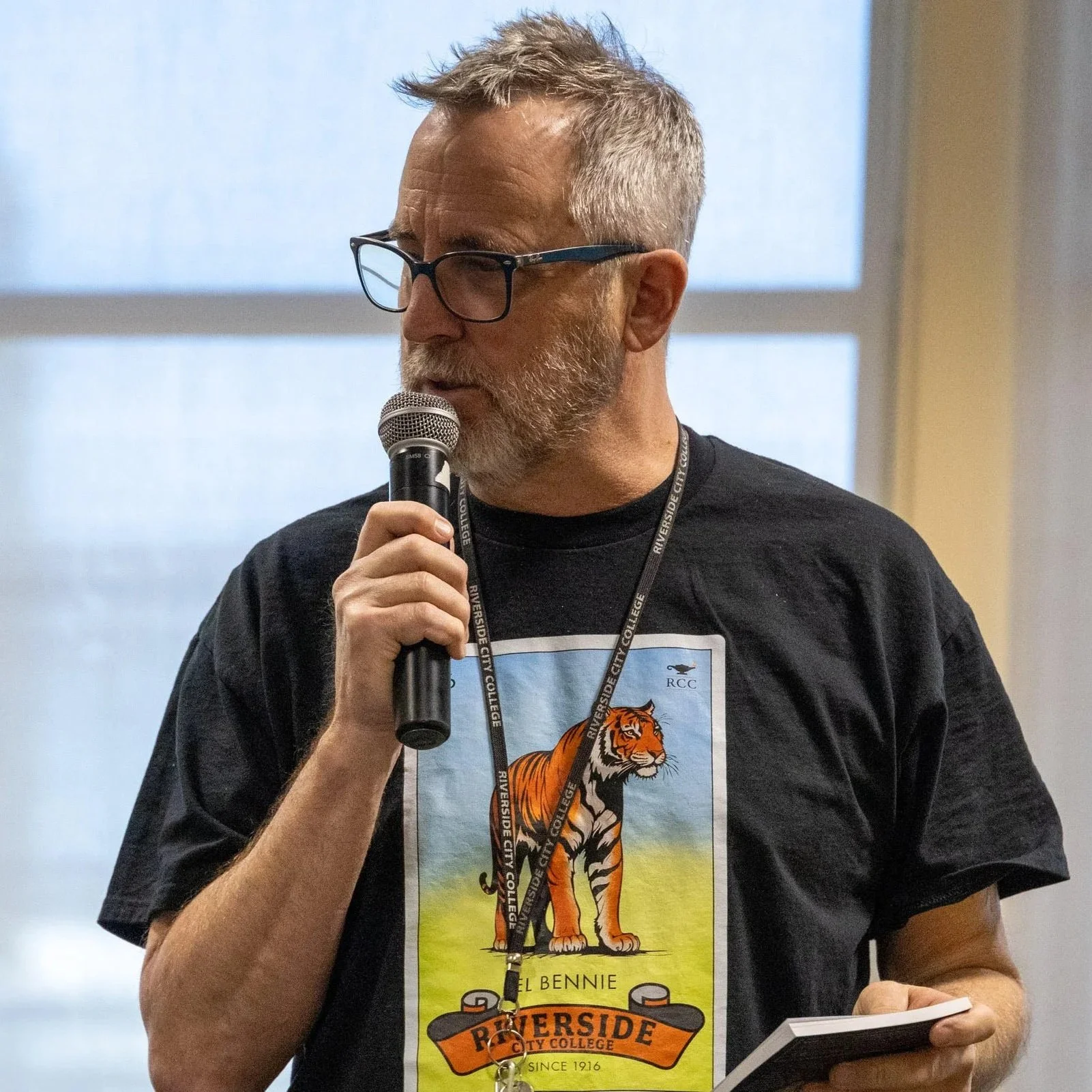 A man with glasses and facial hair speaking into a microphone, wearing a Riversidel City College lanyard and a black t-shirt with a colorful tiger logo and the text 'RIVERSIDE CITY COLLEGE SINCE 1916'.