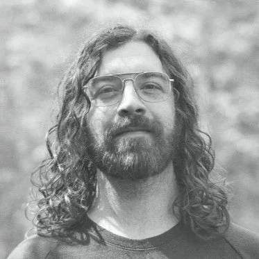 A man with long curly hair, glasses, and a beard, standing outdoors.