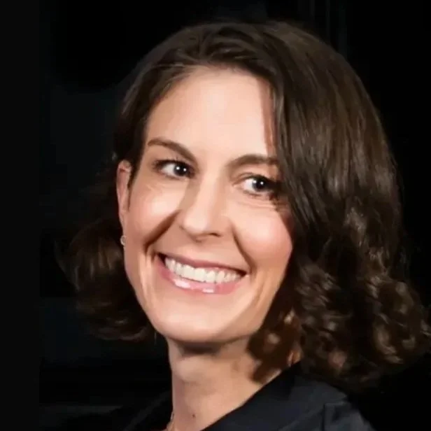 A smiling woman with shoulder-length brown hair and earrings, wearing a dark top, against a dark background.