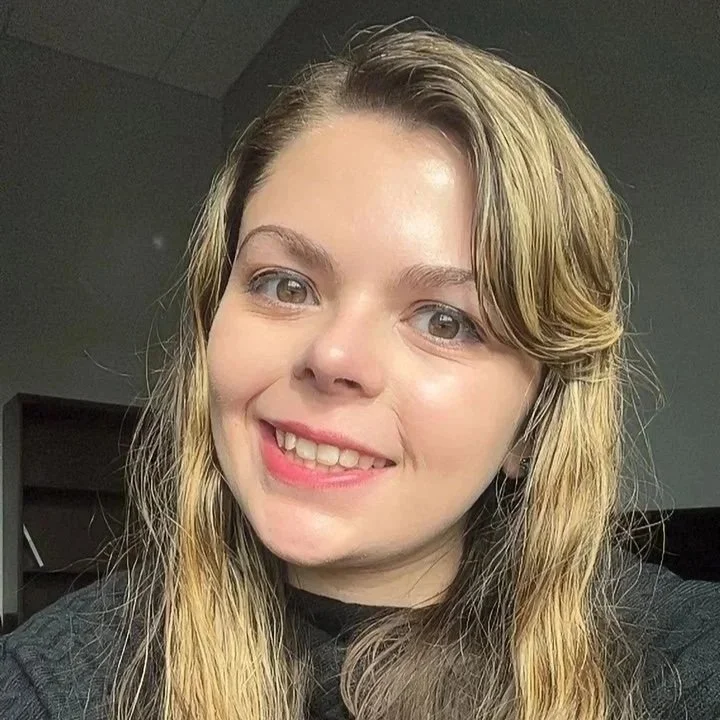 A young woman with long blonde hair, blue eyes, and a friendly smile, wearing a dark top, indoors with neutral background.