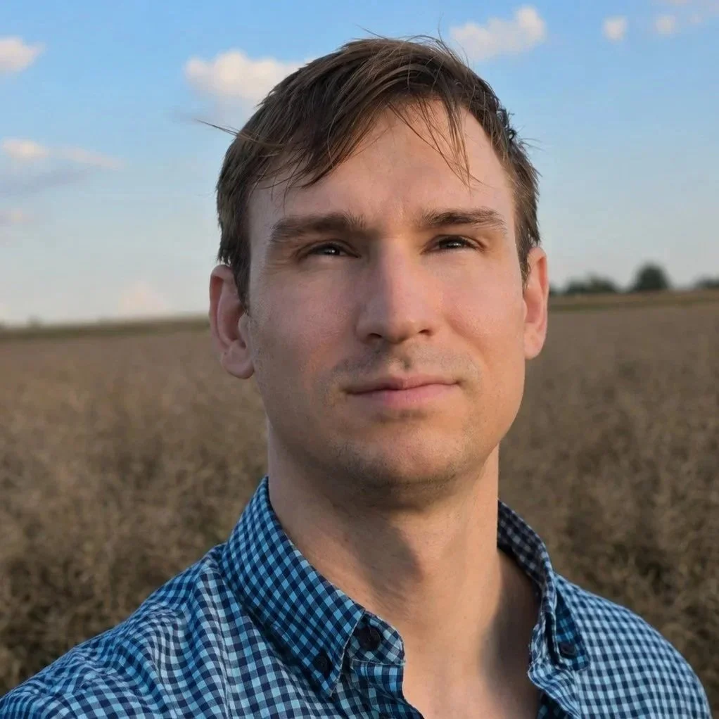 Portrait of a young man with light brown hair, wearing a blue checkered shirt, standing outdoors in a field with a partly cloudy sky.