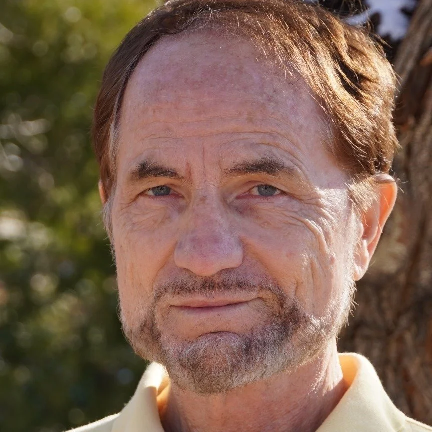 Close-up of a middle-aged man with light brown hair, blue eyes, a beard, and a mustache outdoors near a tree, with sunlight on his face.