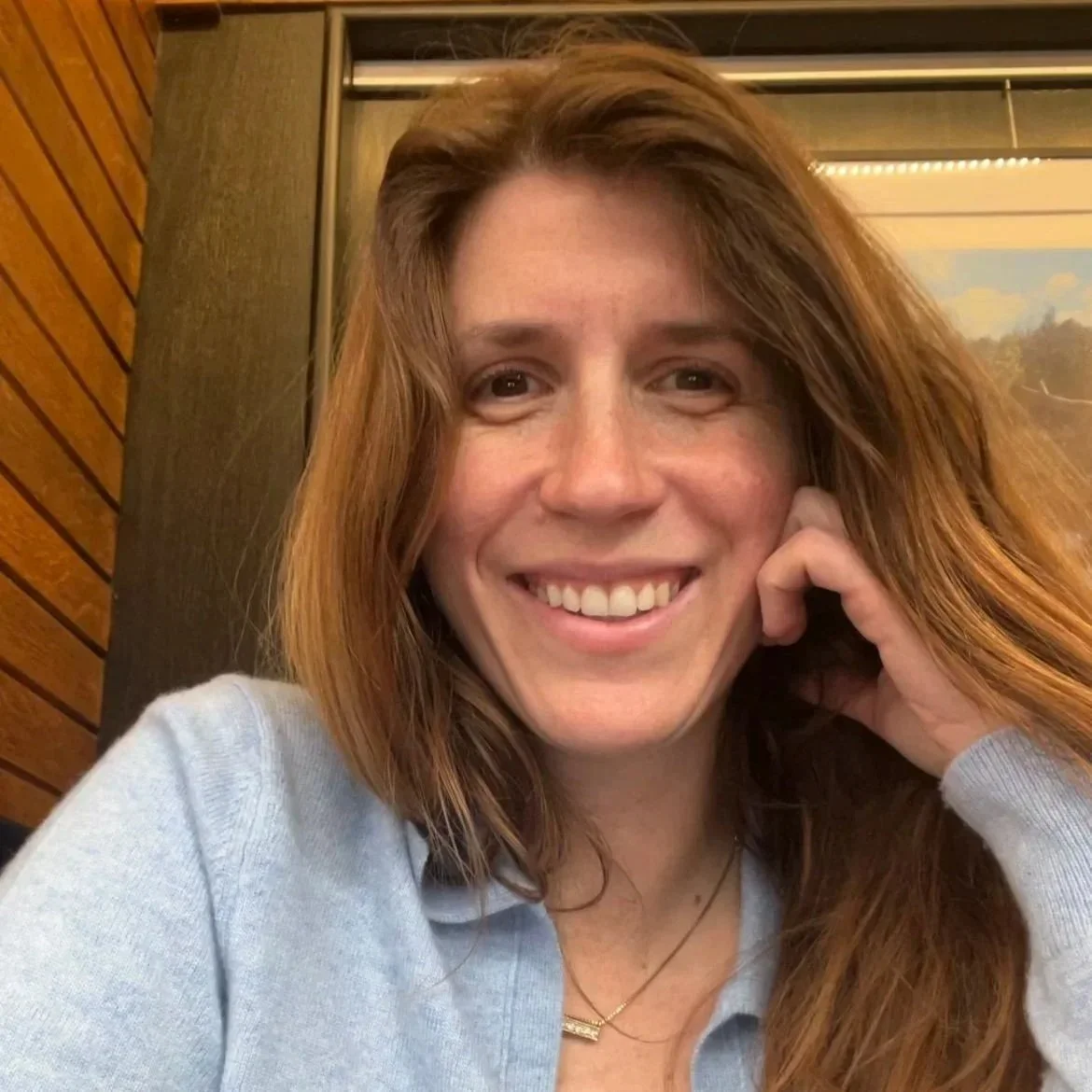 Close-up of a smiling woman with long red hair, wearing a light blue shirt in an indoor setting with wooden paneling and a framed landscape picture in the background.
