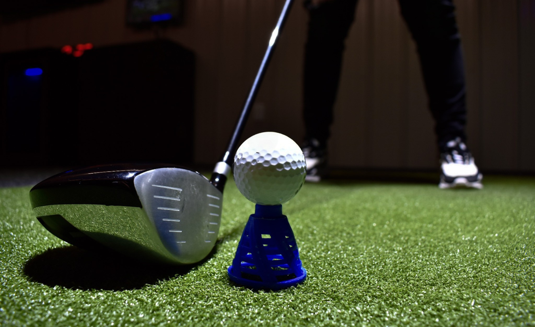 Close-up of a golf driver club and a golf ball on a tee on artificial turf, with a golfer in the background preparing to swing.