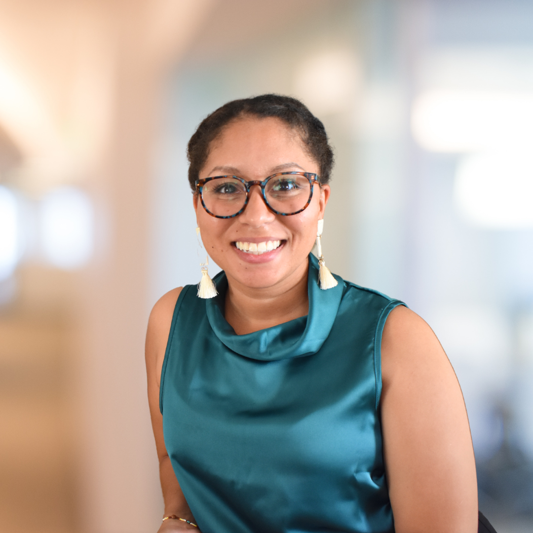 A woman with short curly hair, glasses, and tassel earrings, smiling and wearing a teal satin sleeveless dress.