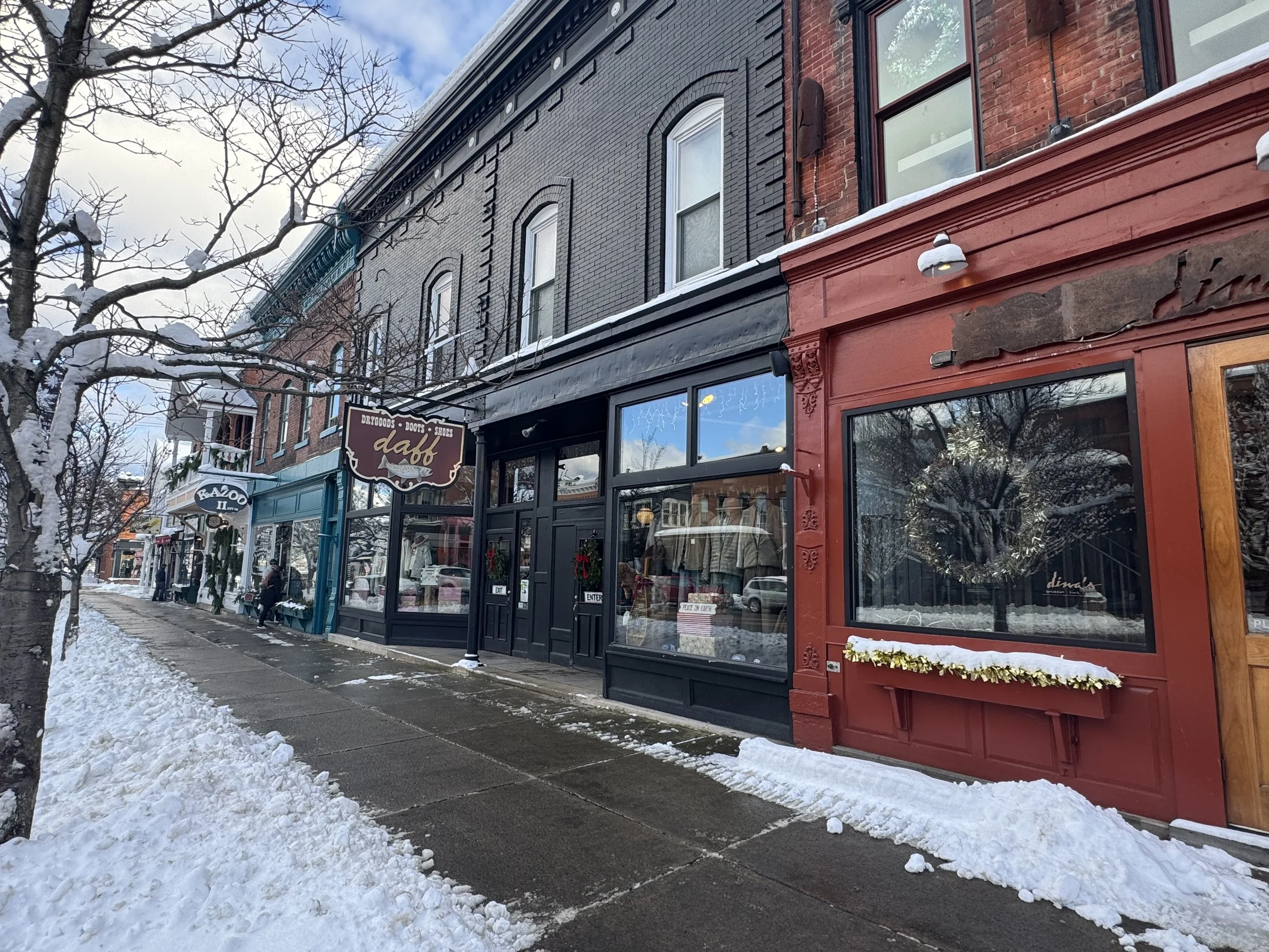 A row of storefronts on a snowy sidewalk, with signs for a dry goods and boots store named 'dahl' and another store with a sign partially visible, in a historic brick building with large windows and awnings.