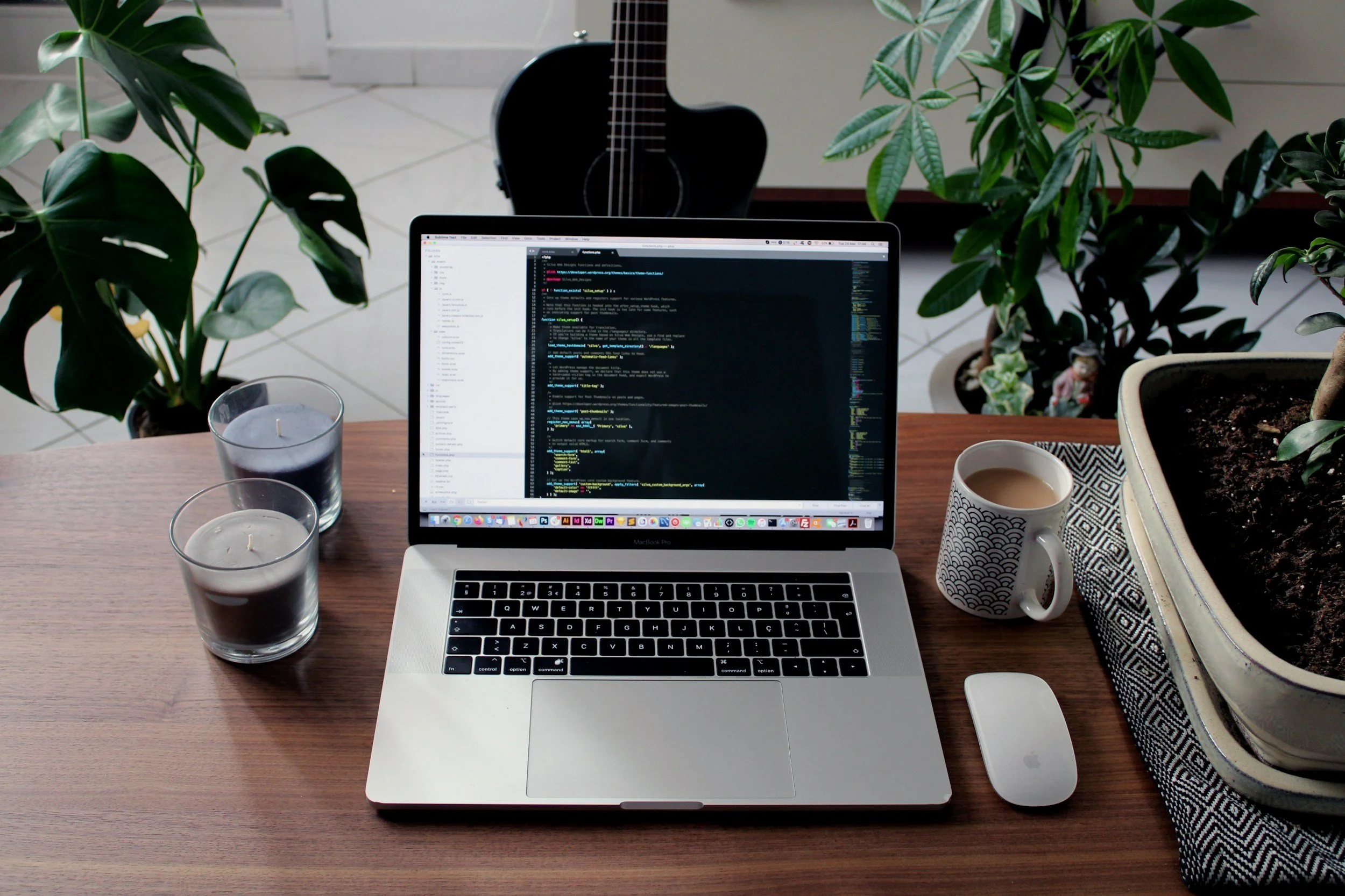 A workspace with a laptop displaying code, two candles, a coffee mug, a mouse, and a keyboard on a wooden desk, surrounded by houseplants and a guitar in the background.