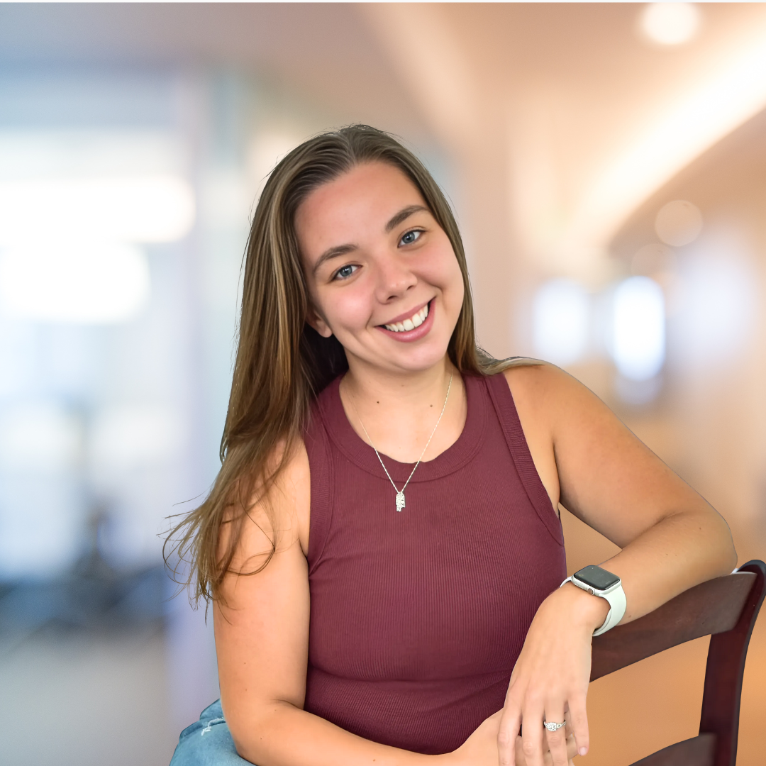 A young woman with long brown hair smiling, wearing a maroon sleeveless top, a silver necklace, a smartwatch, and a ring, sitting at a table in a brightly lit indoor setting.