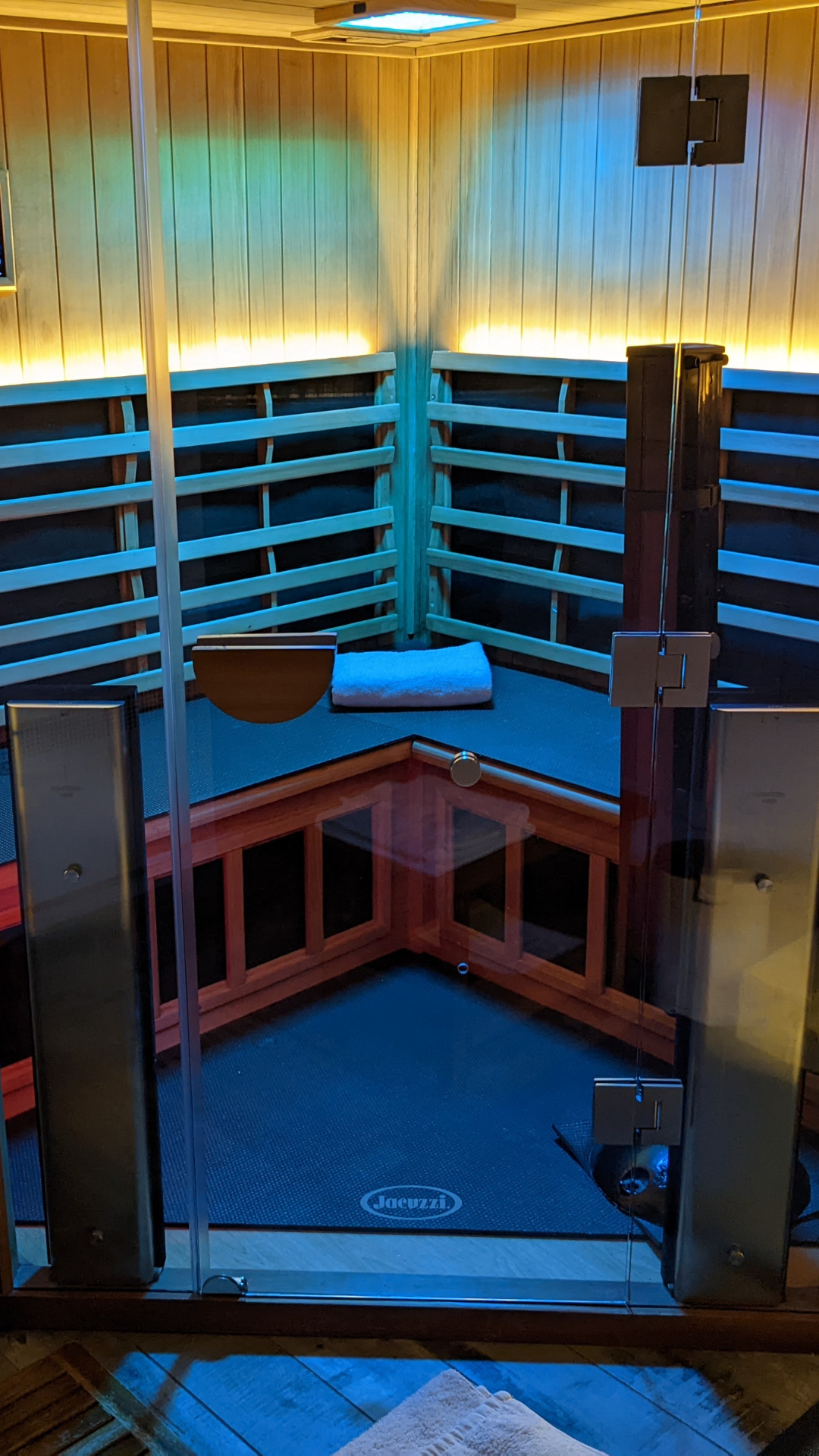 A steam or infrared sauna room with wooden walls and shelving, a white rolled towel, a wooden bucket, and a Jacuzzi branded floor mat, viewed through a glass door.