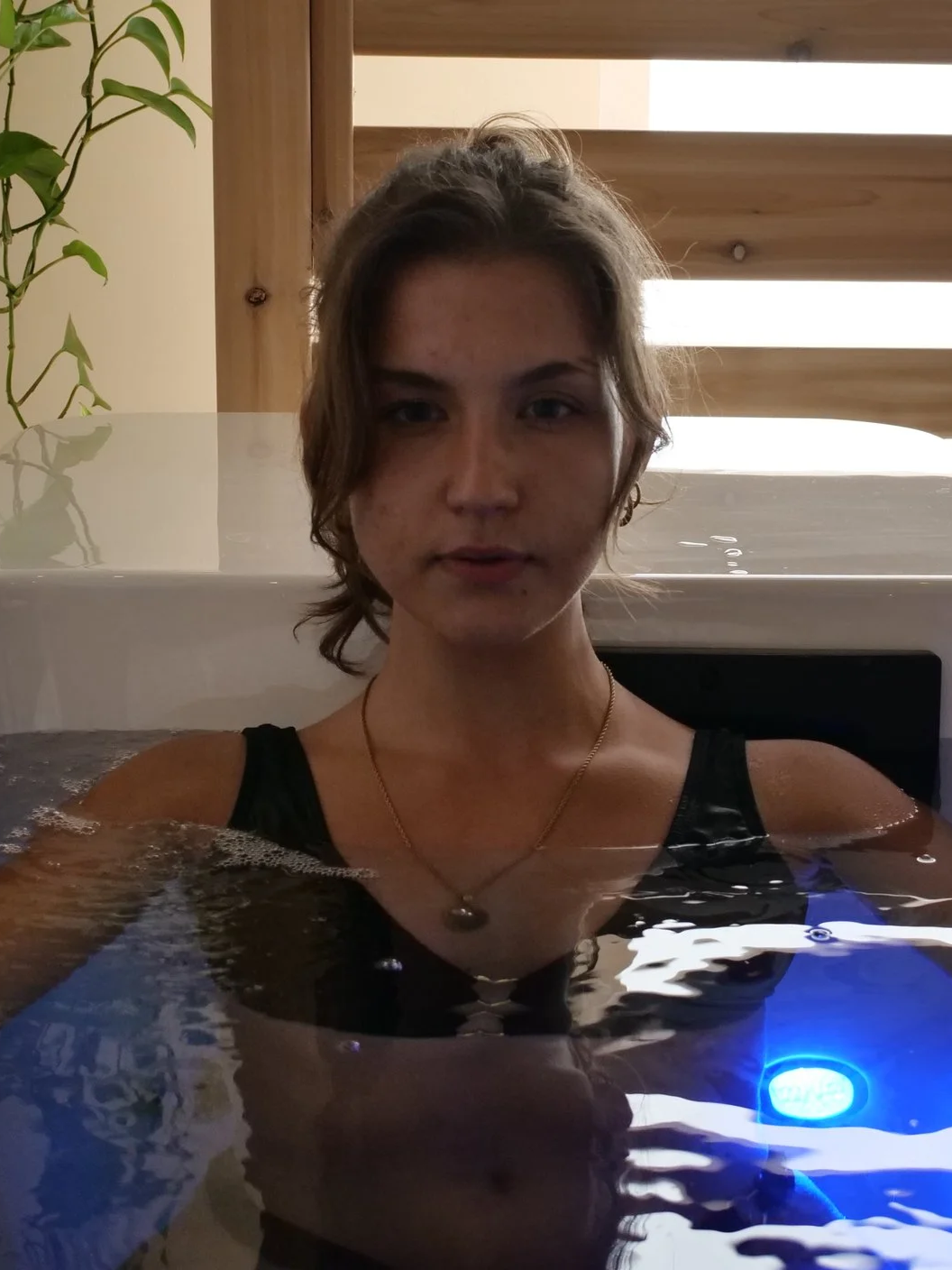 A young woman with short, wavy brown hair taking a bath in a hot tub indoors. She is wearing a black top and a gold necklace with a pendant, with wood paneling and a plant in the background.