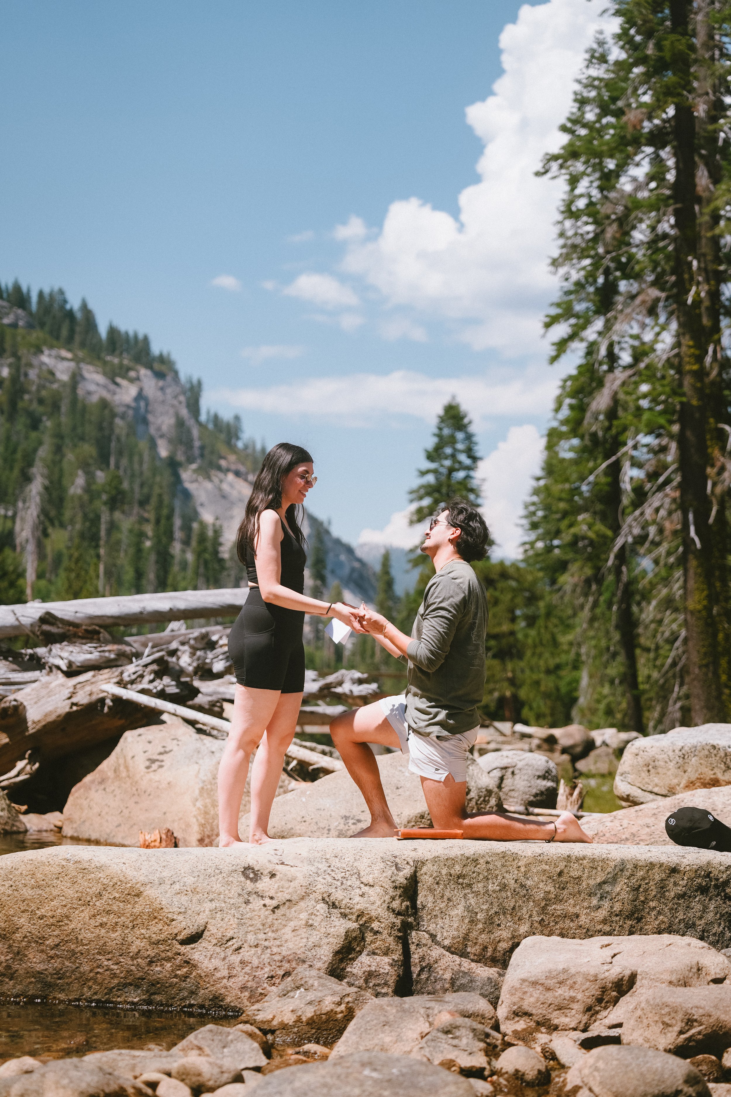 A couple getting engaged by a river in a forested mountain area on a sunny day, with the man down on one knee holding the woman's hand.