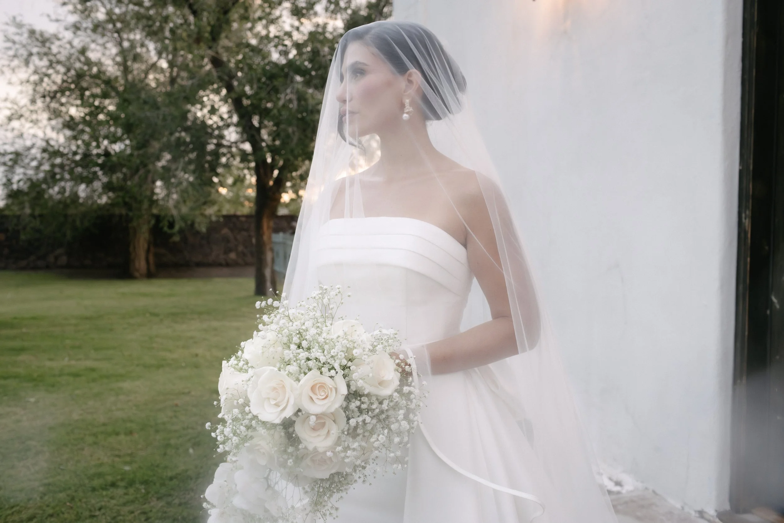 A bride in a white wedding dress holding a bouquet of white roses and baby's breath, standing outdoors near a white wall with trees in the background, wearing a veil and pearl earrings. Wedding outdoors. bride. white dress. flowers.