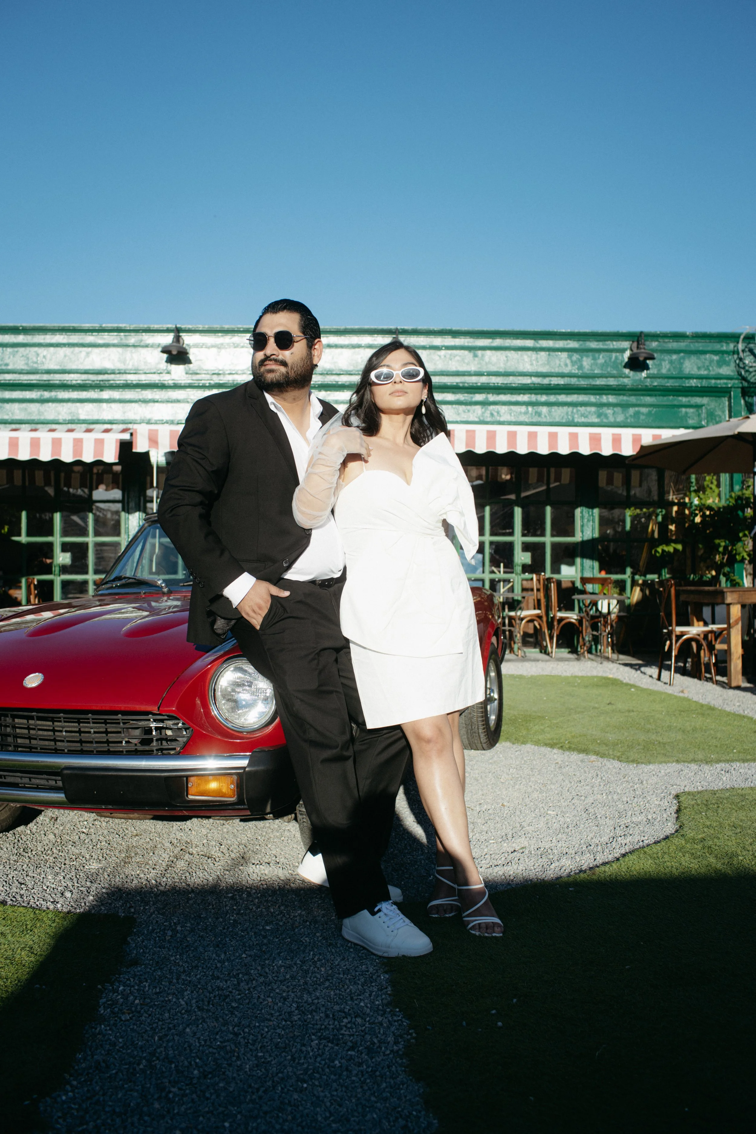 A man and woman dressed in stylish fashion posing beside a red vintage car outdoors, with a green building and outdoor seating area in the background. The man is wearing a black suit, white shirt, and sunglasses, while the woman is in a white dress and sunglasses.