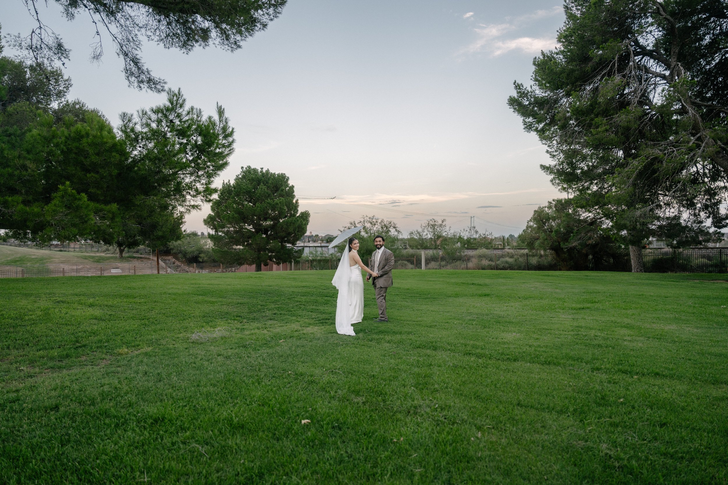 A bride and groom holding hands standing on a grassy field with trees and a fence in the background, under an open sky at dusk.