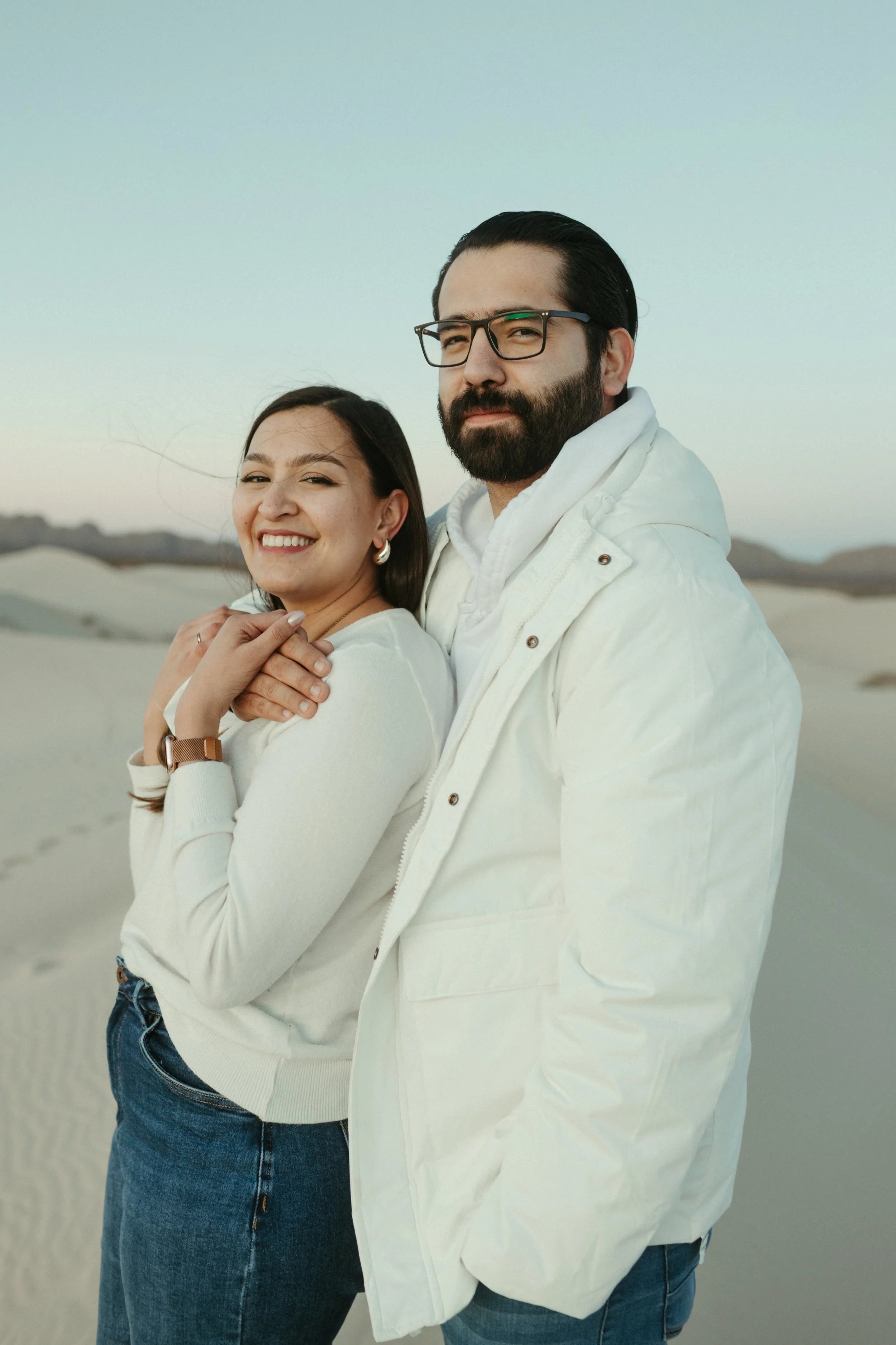 A couple standing in a desert landscape, both wearing white jackets. The woman is smiling and has her hands crossed over her chest, while the man has a serious expression and wears glasses.