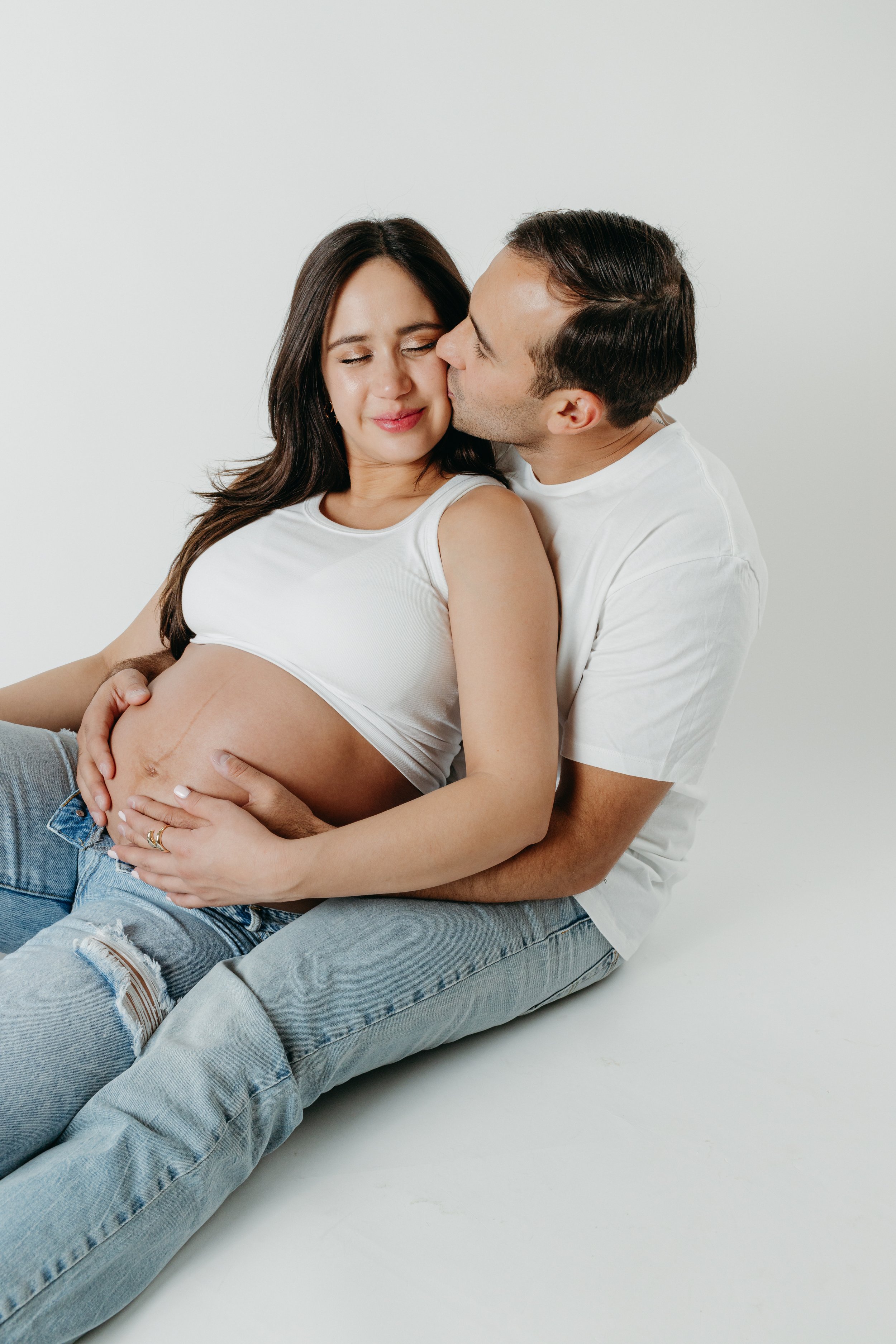 Pregnant woman with long dark hair sitting on the floor with her partner, who is kissing her cheek, while her hand rests on her belly.