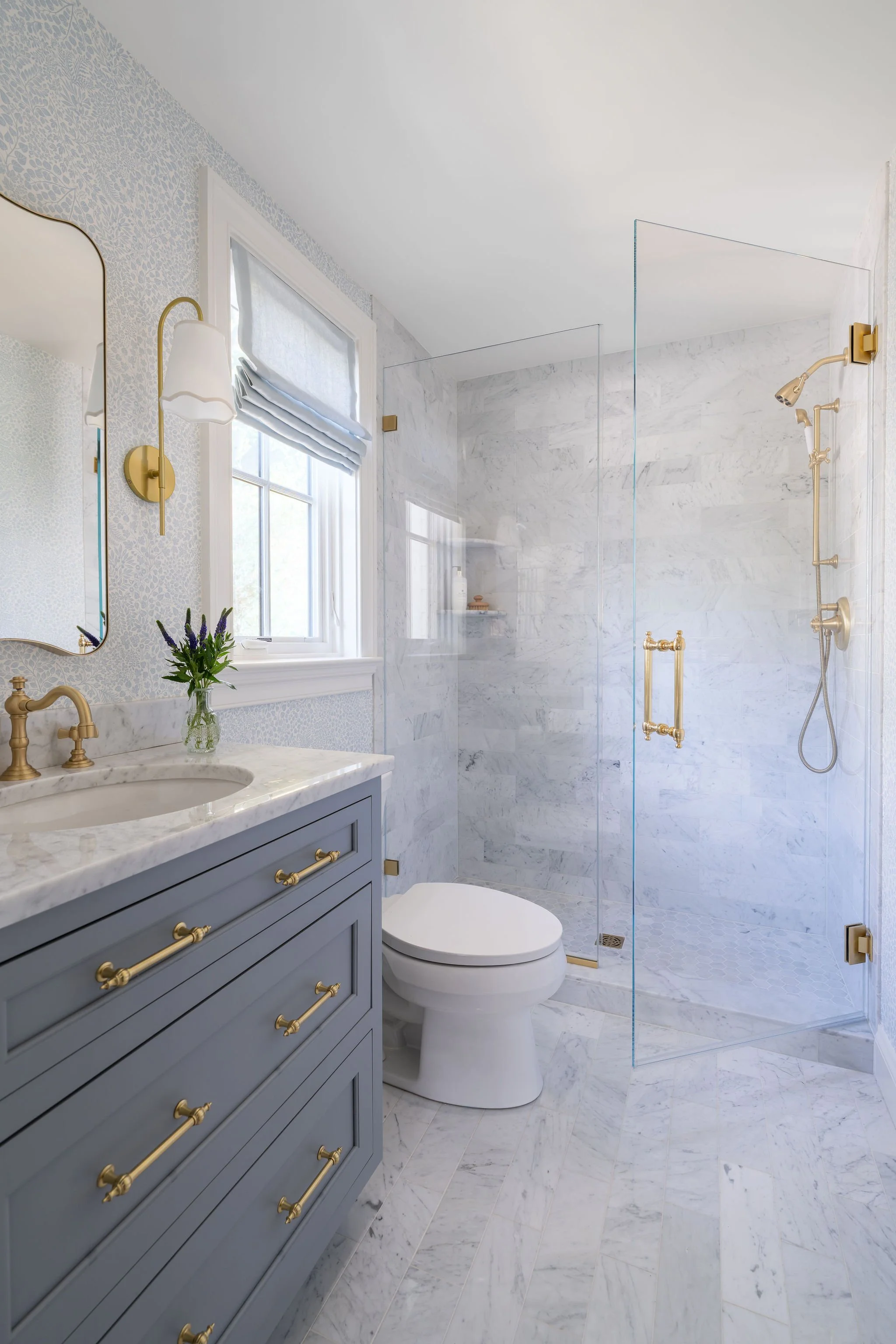 A marble bathroom with coastal blues, petite print wallpaper, brass accents, and custom roman shades. 
