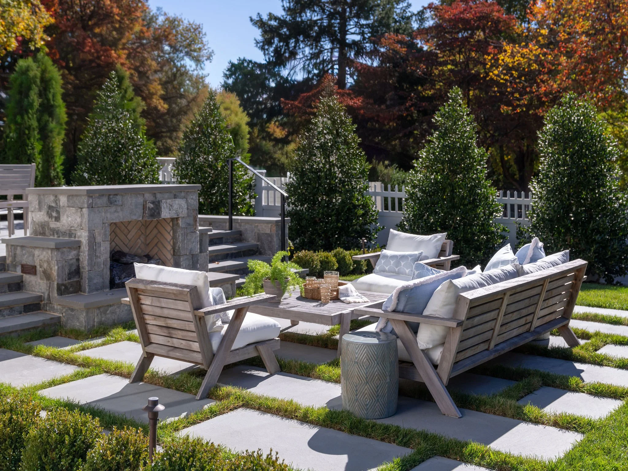 An outdoor entertainment space with teak lounge furniture, a masonry fireplace and pavers set in lush green grass. 
