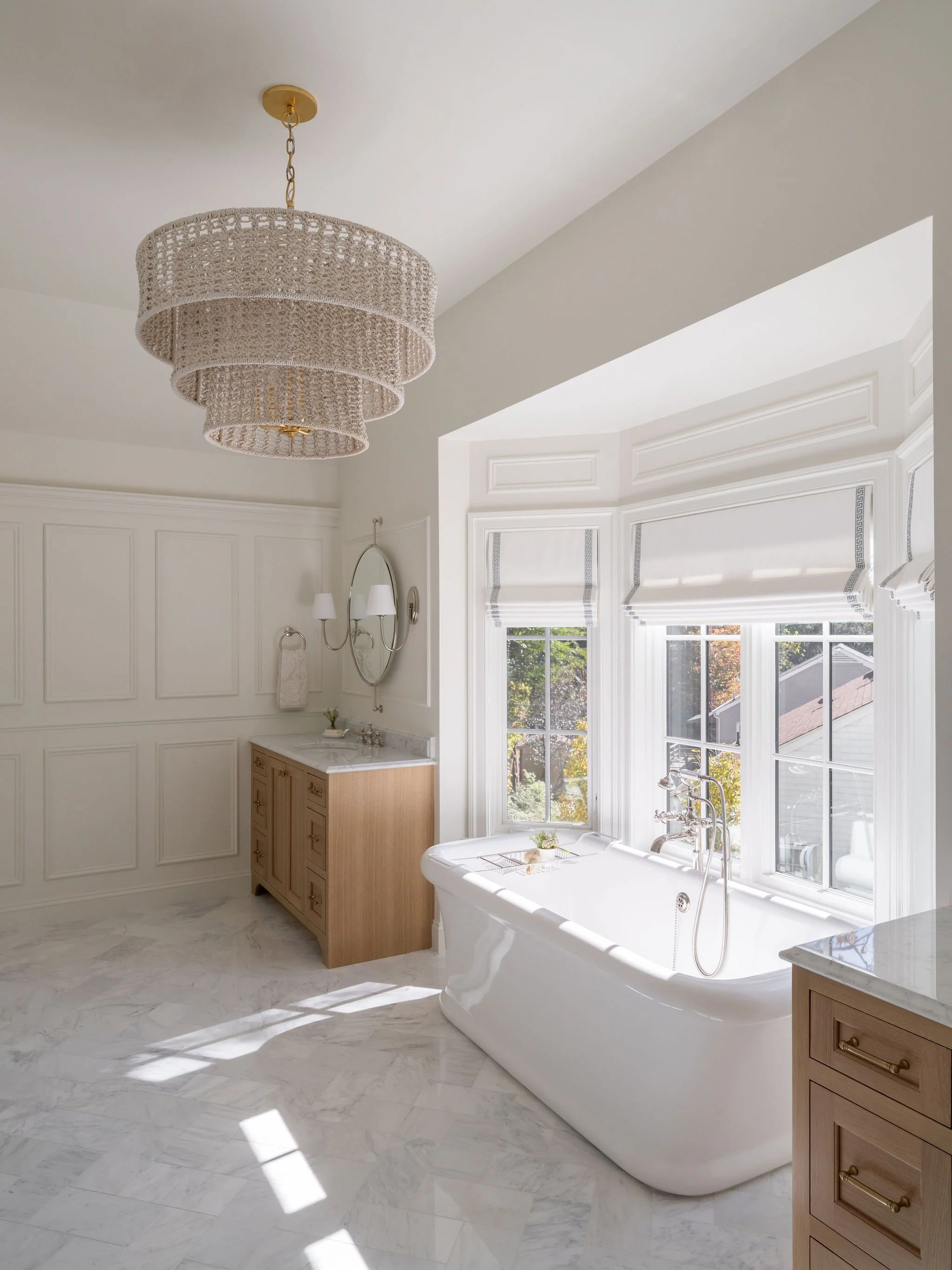 A soaking tub set in a bay window nook with custom roman shades, flanked by oak vanity cabinets and accented by a tiered chandelier. 