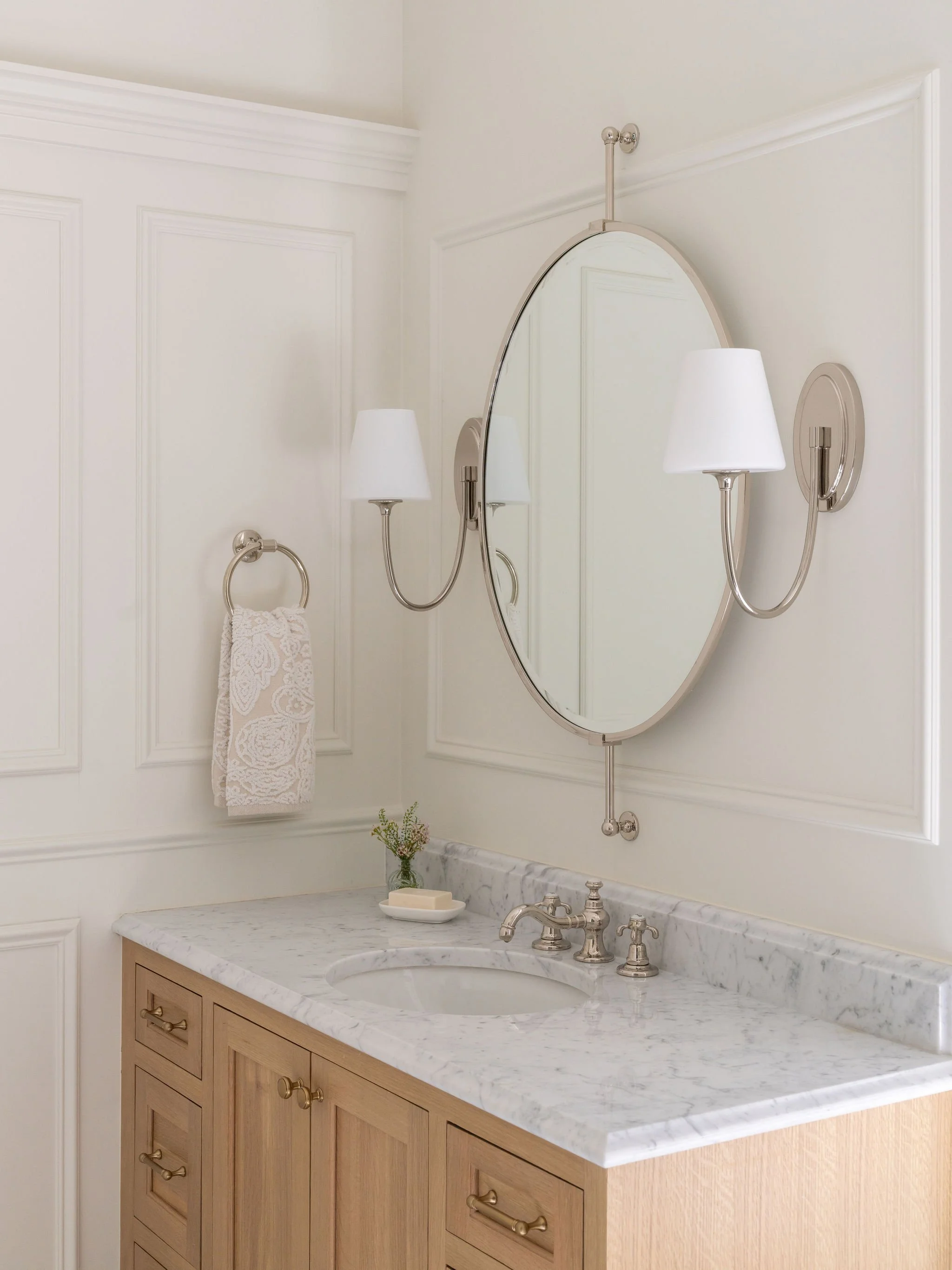 A white oak vanity with marble countertops and a polished nickel oval mirror and shaded sconces. 