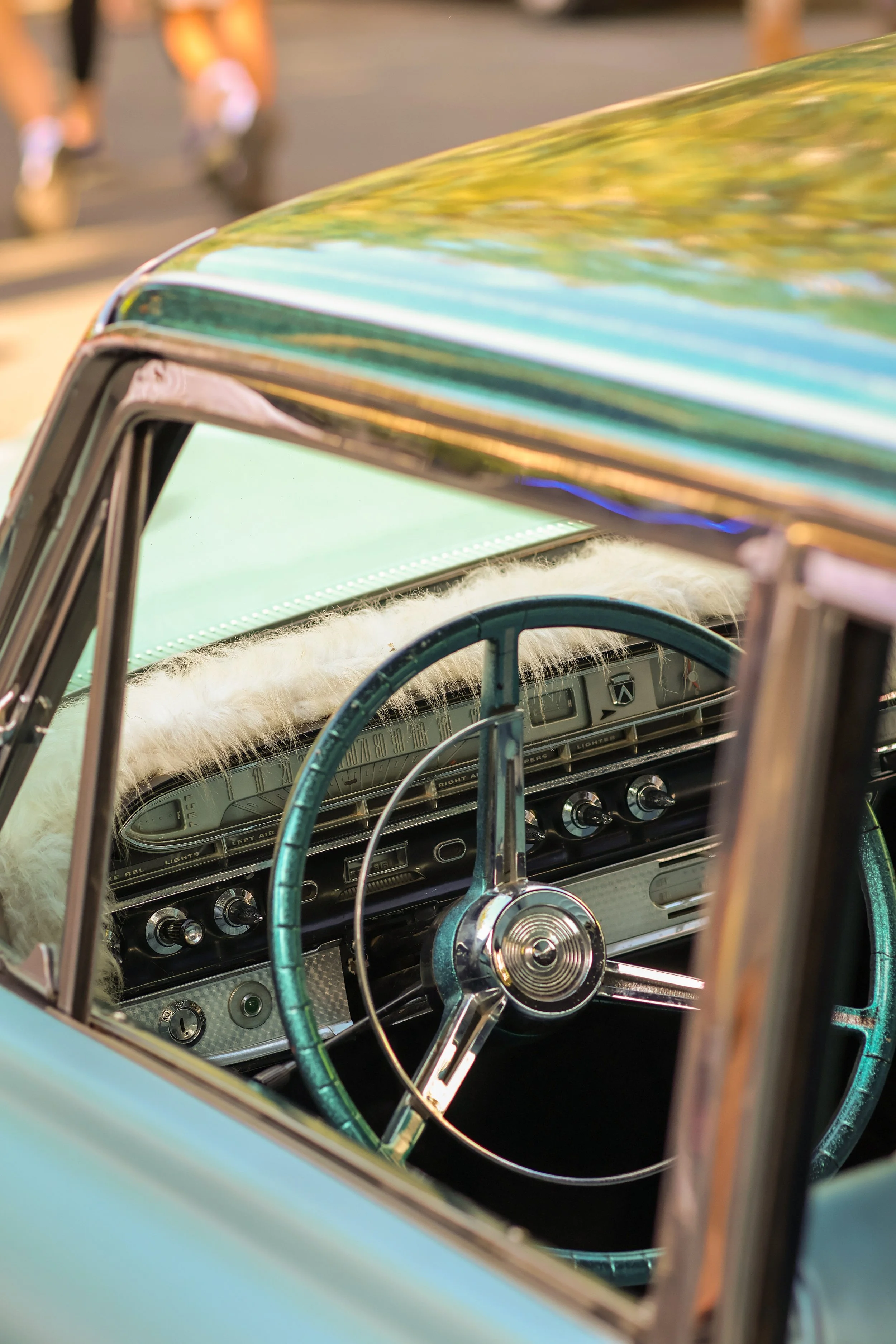 Close-up of the interior of a vintage turquoise car, showing the dashboard, steering wheel, and part of the windshield, with people walking in the blurred background.