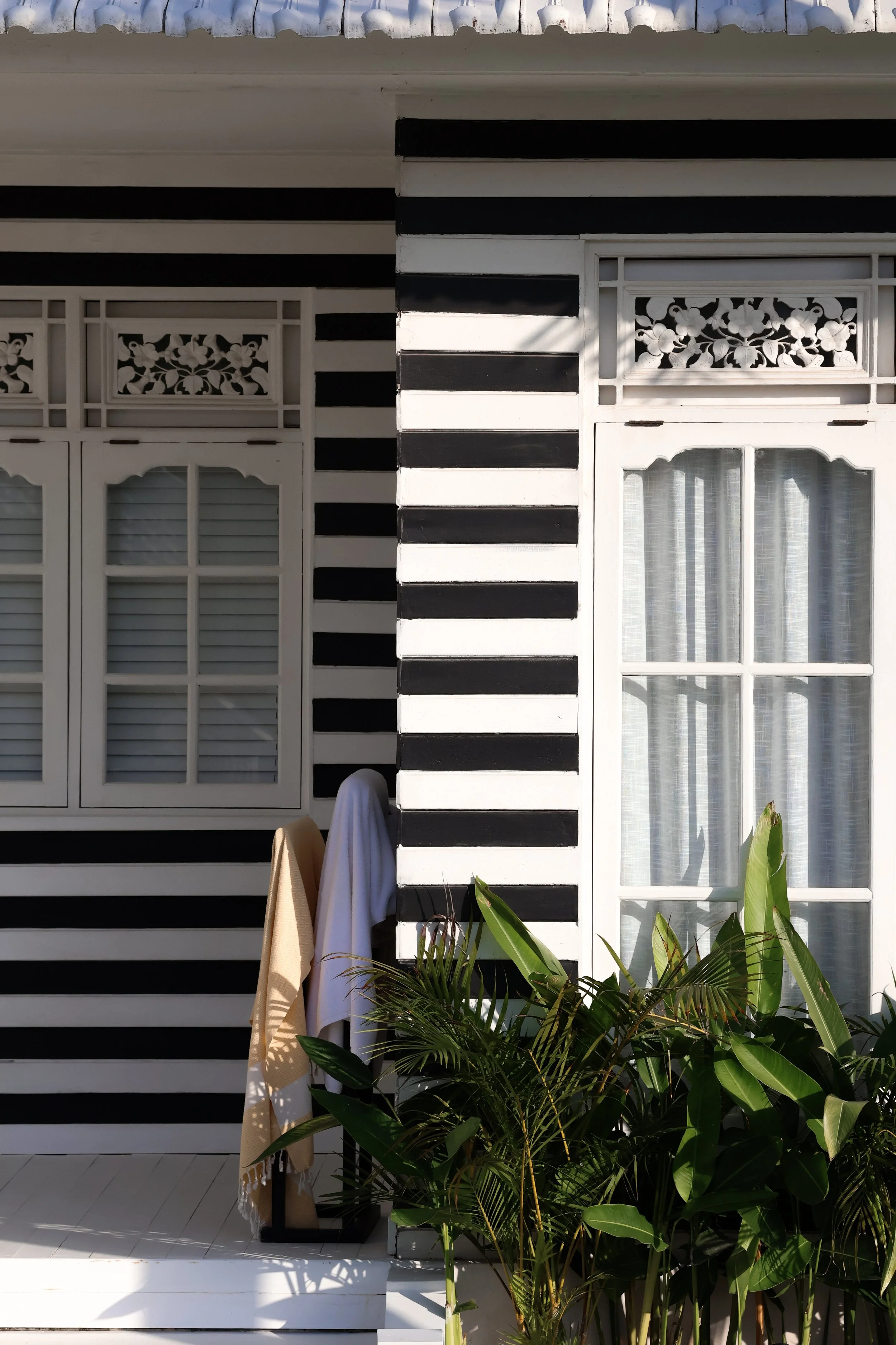 Exterior wall of a building painted with horizontal black and white stripes, featuring two white framed windows with floral and paneled details, surrounded by green plants.