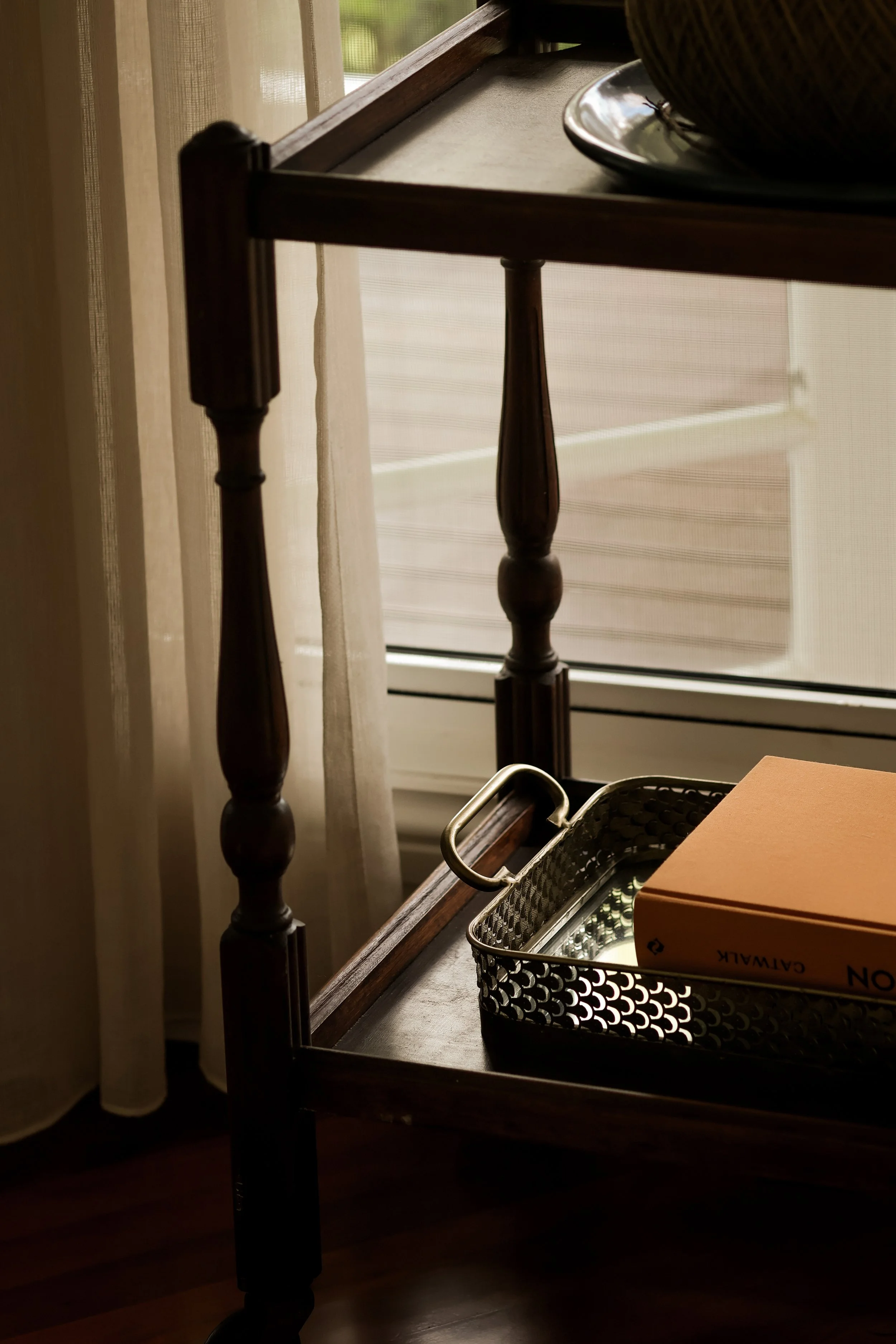 A wooden side table near a window with cream-colored curtains. On the table, there is a metal tray with a yellow box labeled 'CARAMEL' and some other items. In the background, outdoors greenery is visible through a window screen.